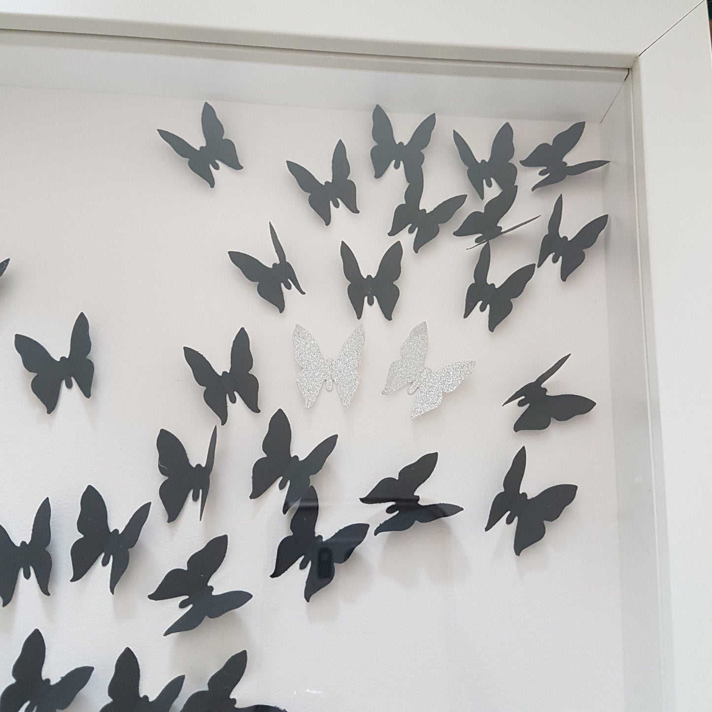Square white box frame showing a flurry of 3D black paper butterflies with two silver glitter butterflies, moving from the bottom left of the frame, through to the top right of the frame. Shown against a white background