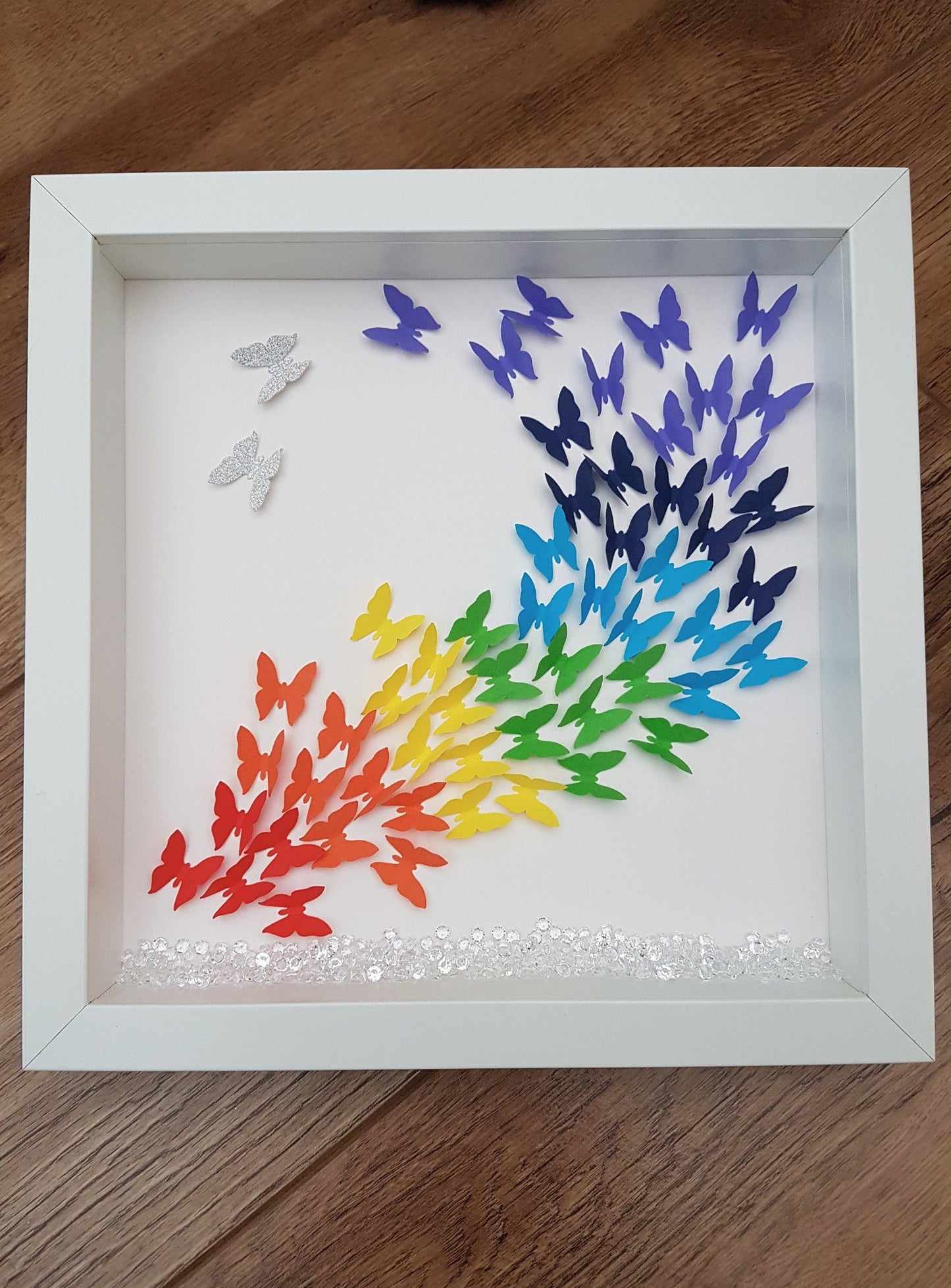 Square white box frame showing a flurry of paper butterflies in rainbow colours, moving from red butterflies in the bottom left of the frame, through rainbow colours to the top right of the frame. Shown against a wooden background