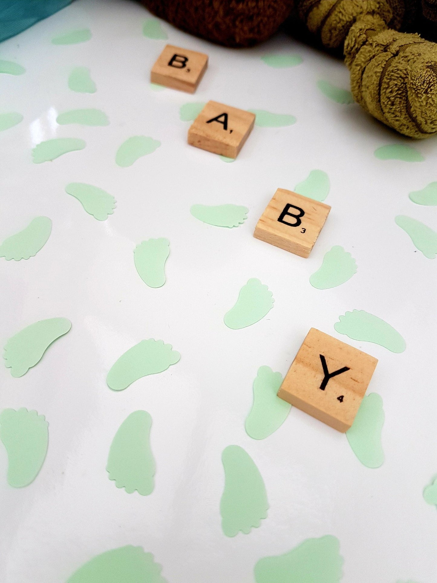 Pastel mint green baby feet shaped confetti pieces are scattered on a white background with wooden scrabble tiles reading BABY diagonally across the photo. Cuddly toys are seen in the background