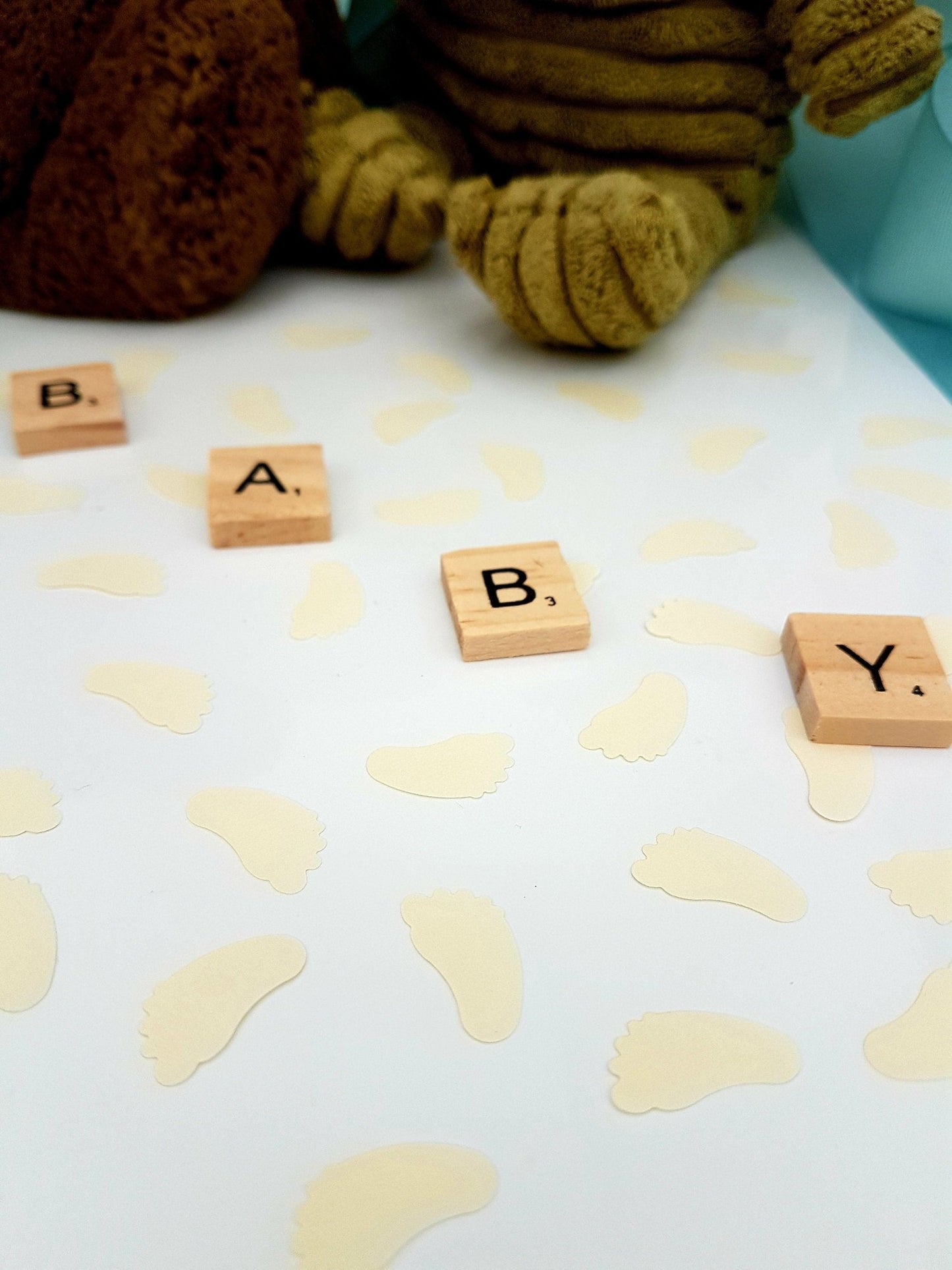 Cream ivory baby feet shaped confetti pieces are scattered on a white background with wooden scrabble tiles reading BABY diagonally across the photo. Cuddly toys are seen in the background