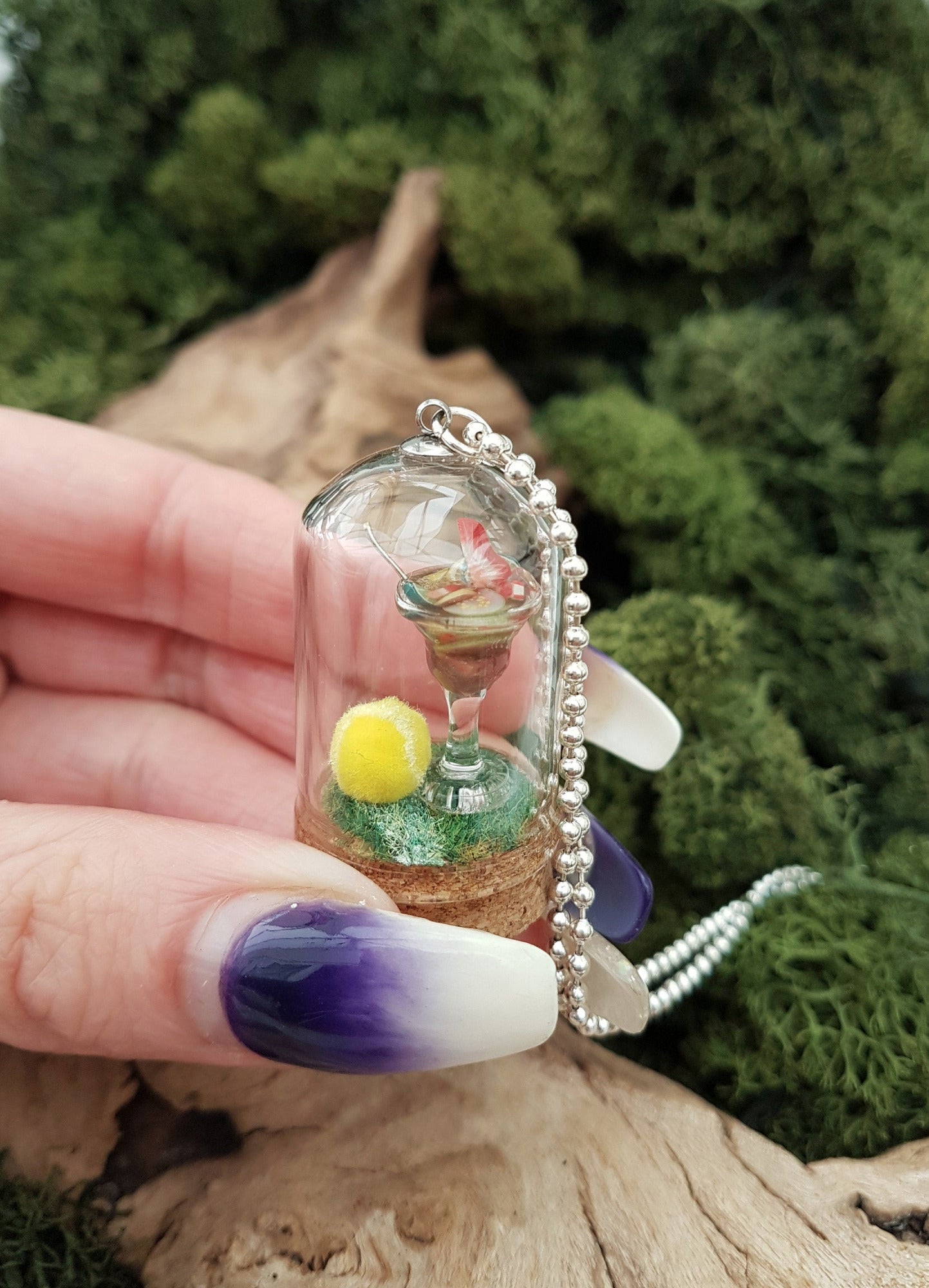Miniature glass bell jar containing a cocktail glass with Pimm's and miniature fruit slices sat on a green flocked base with a fluorescent yellow tennis ball. Held between finger and thumb. A silver coloured chain is attached to the top