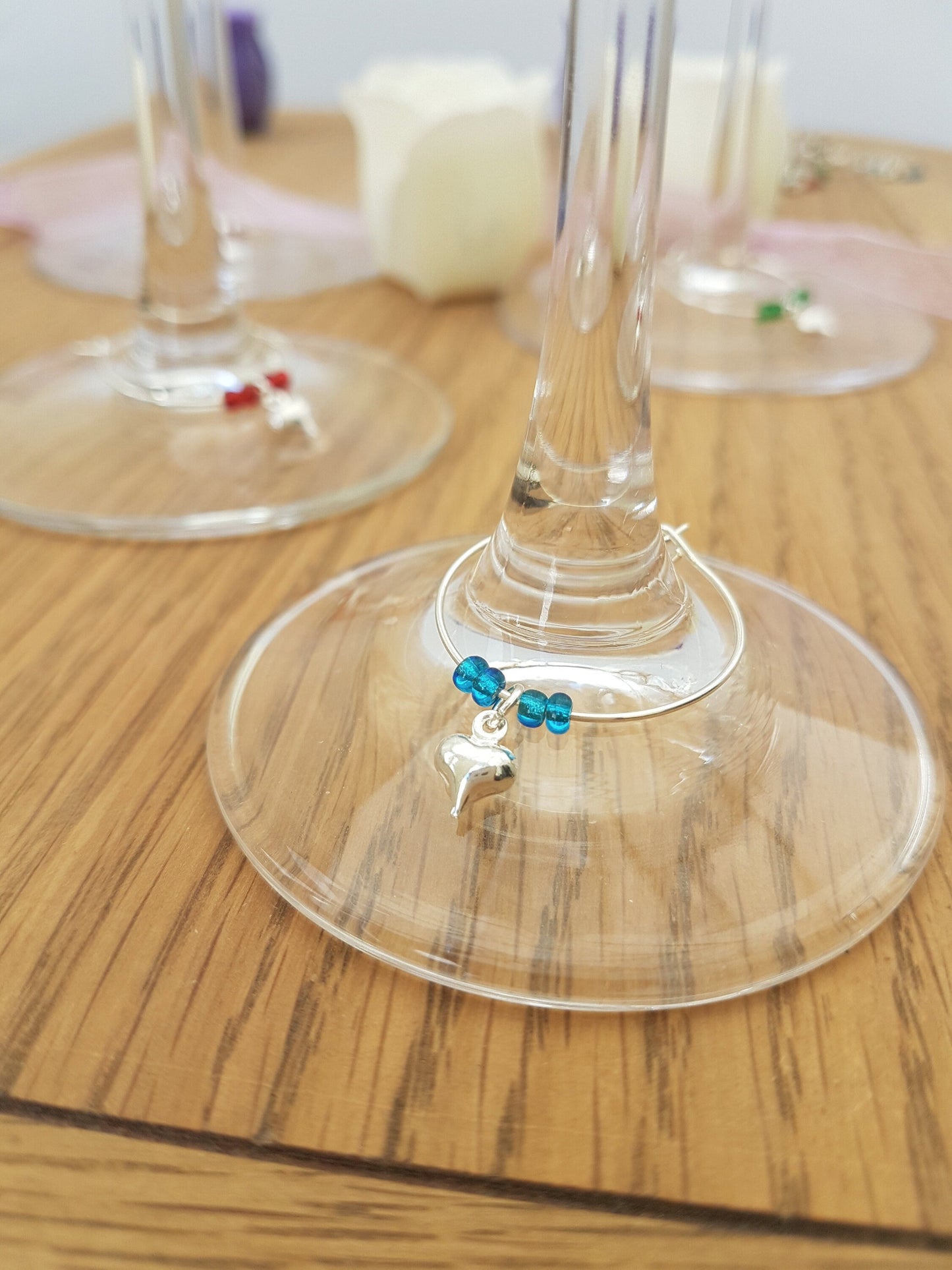 3 wine glass charms are attached to 3 champagne flute bases on a wooden background. The wine glass charms are round wire hoops, each with different coloured seed beads and a silver coloured puffy heart. The turquoise wine charm is shown in foreground