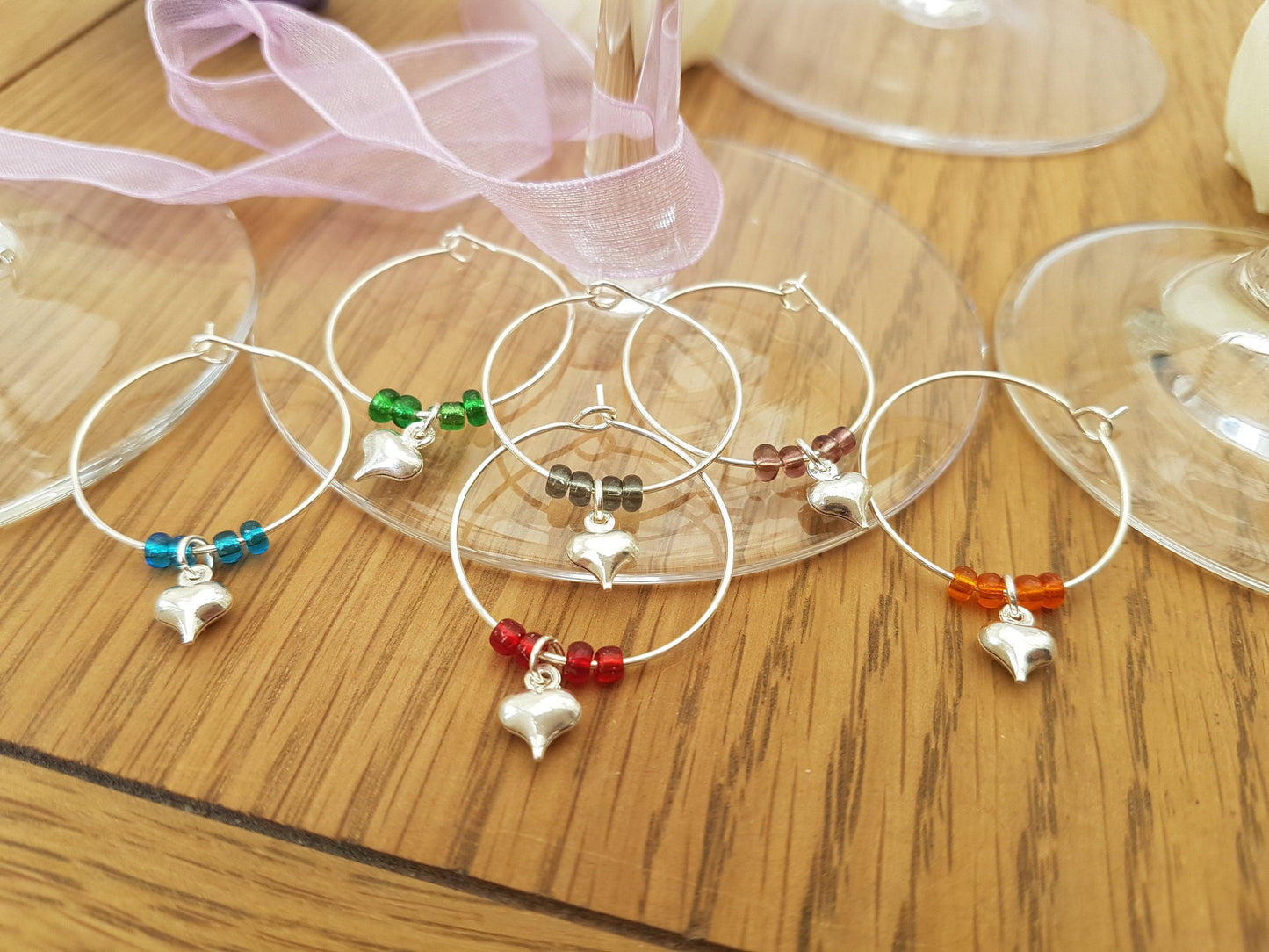 6 wine glass charms are arranged in 2 rows of 3 on top of champagne flute bases on a wooden background. The wine glass charms are round wire hoops, each with different coloured seed beads and a silver coloured puffy heart