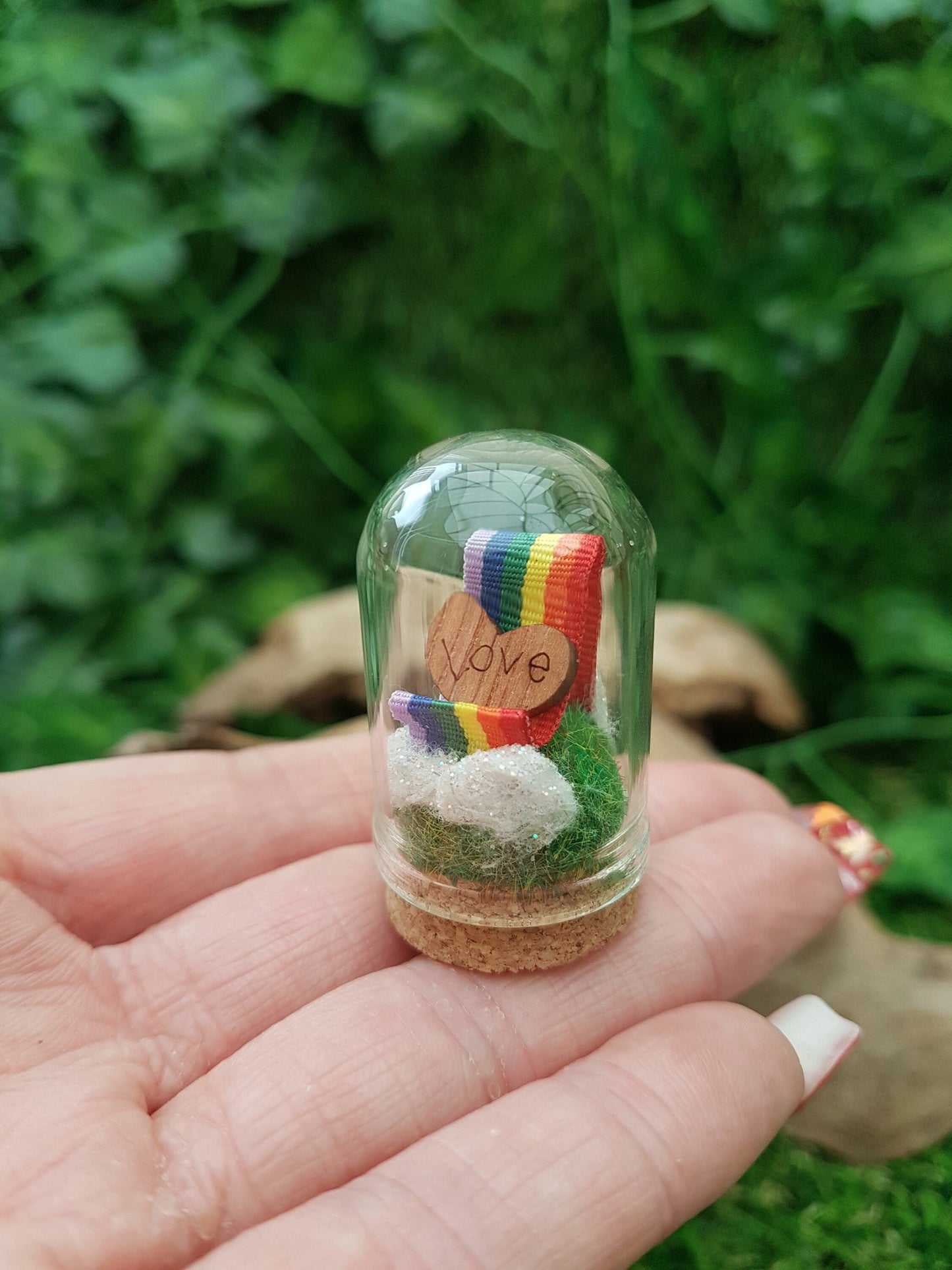 Miniature glass bell jar containing a curl of rainbow ribbon with miniature clouds at the start and end of the ribbon at the back and front. A small wooden heart with sits on the ribbon, engraved with the word LOVE. Dome is displayed on fingers