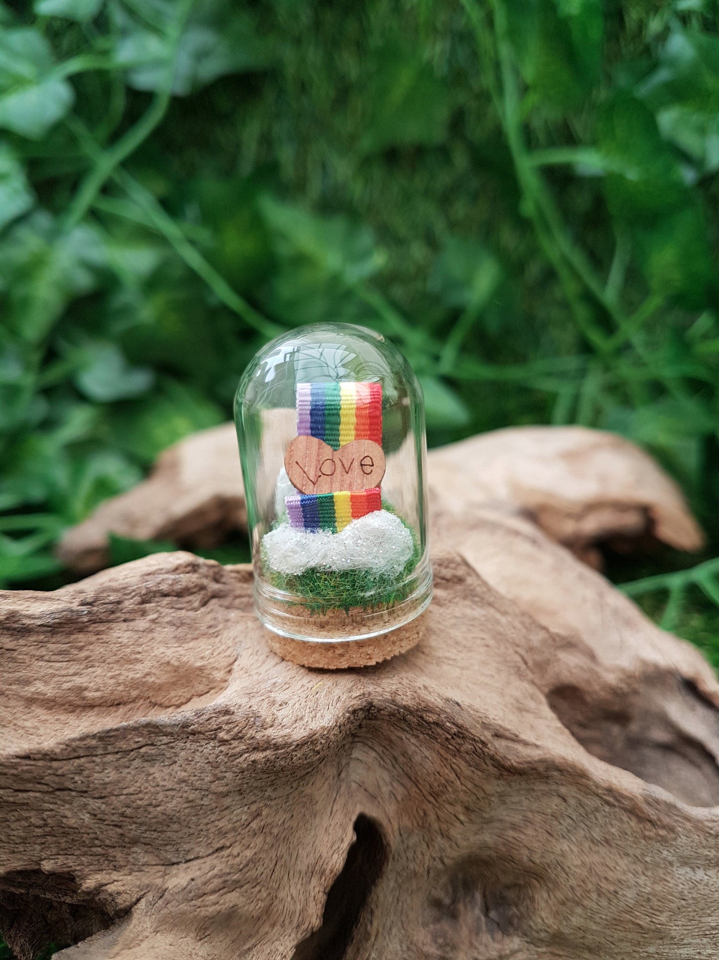 Miniature glass bell jar containing a curl of rainbow ribbon with miniature clouds at the start and end of the ribbon at the back and front. A small wooden heart with sits on the ribbon, engraved with the word LOVE