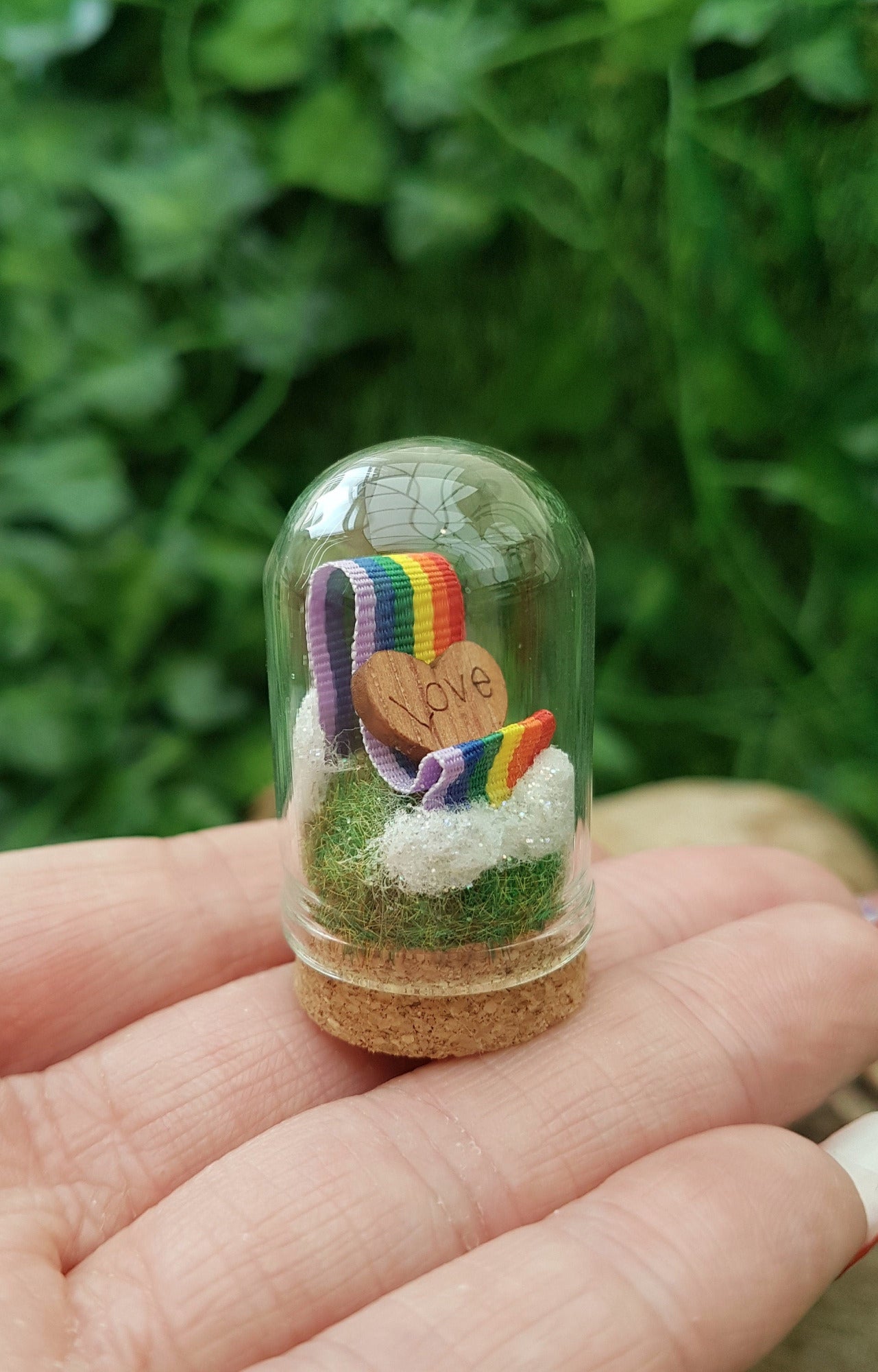 Miniature glass bell jar containing a curl of rainbow ribbon with miniature clouds at the start and end of the ribbon at the back and front. A small wooden heart with sits on the ribbon, engraved with the word LOVE. Dome is displayed on fingers
