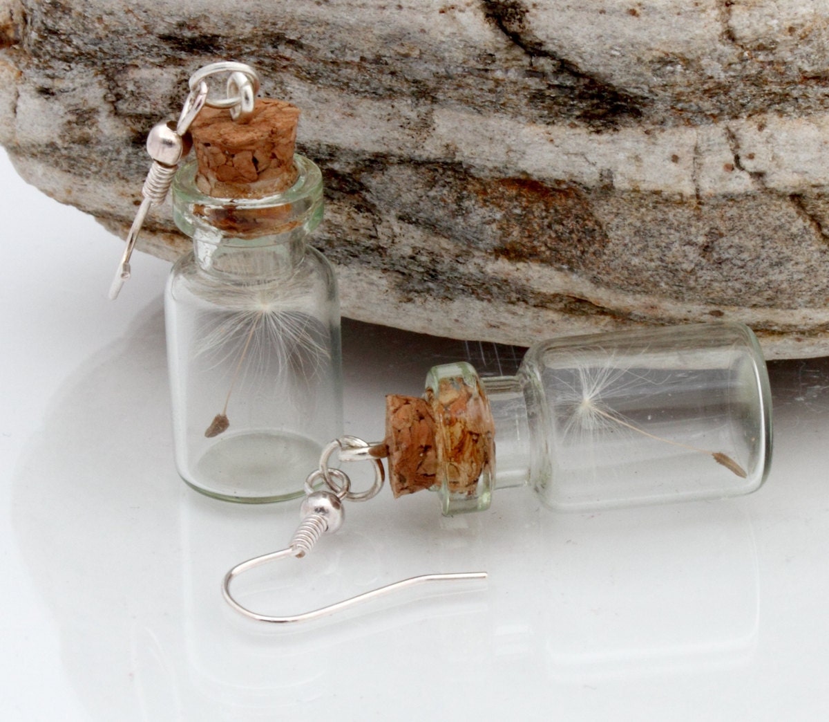 Two small glass jar vials are shown against a natural stone background. One vial is stood up and one vial is in its side, both containing dandelions clock seeds. Each jar has a cork lid with an earring hook attached by jump ring