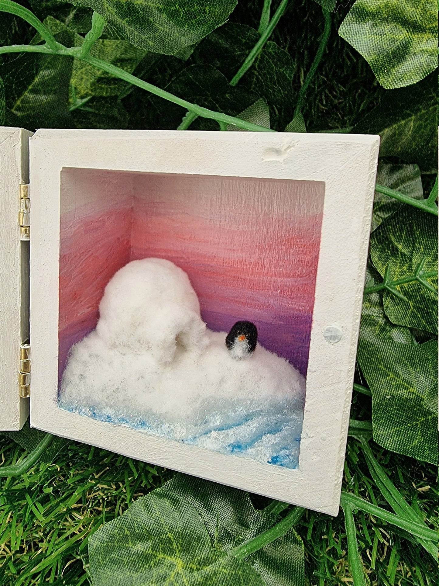 A small white square box, open like a book. Detail is shown of the scene on the right, showing a purple to pink ombre sunset, turquoise and white sea surrounding a snowy iceberg with a white igloo and a tiny penguin.