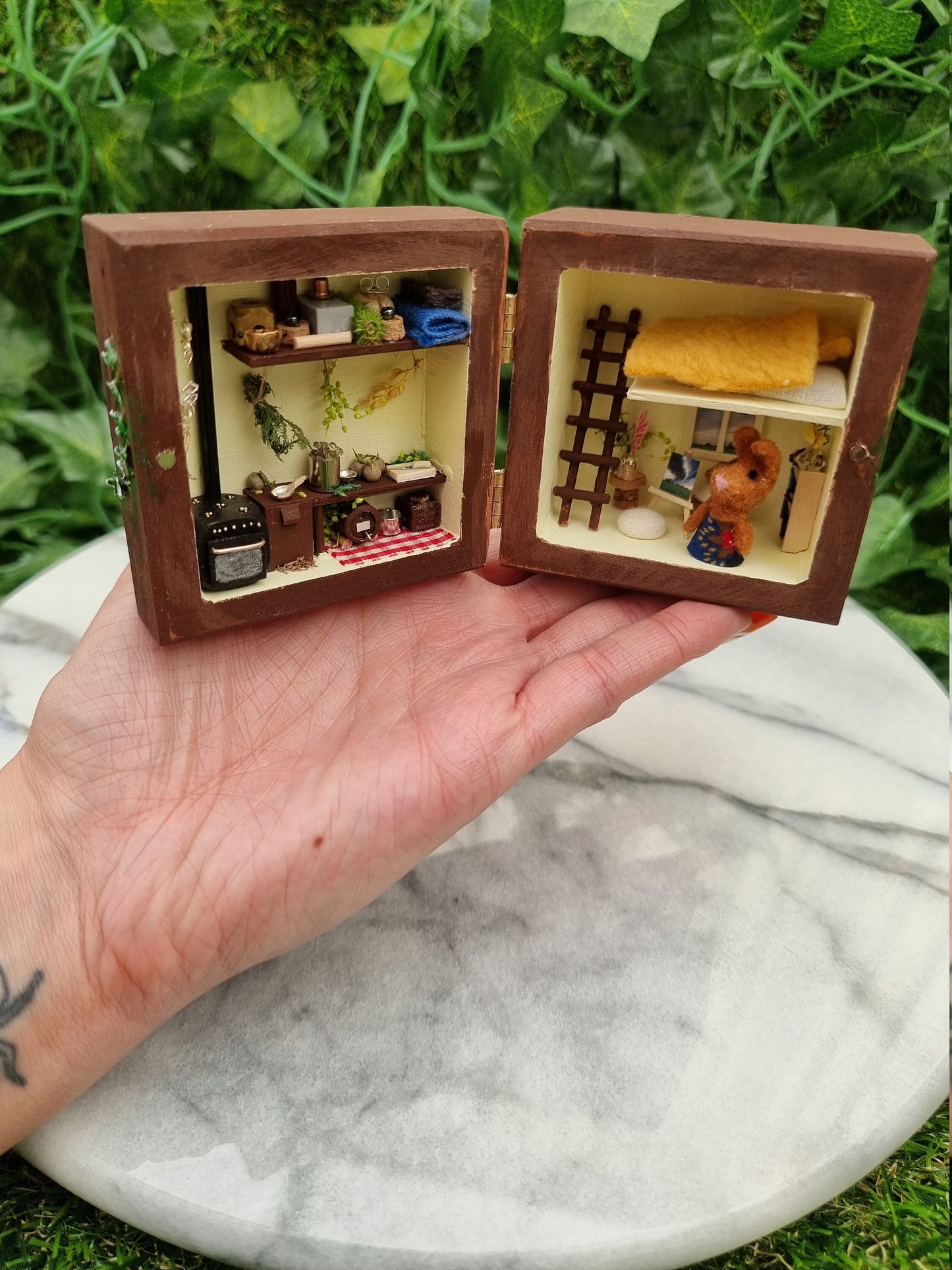 A small brown box sits open like a book on a palm. A small brown bunny in a dress sits in the right side of the box along with a bookcase, easel and ladder to a loft bed. The left side has a tiny oven, table, keg, herbs, tools and storage.