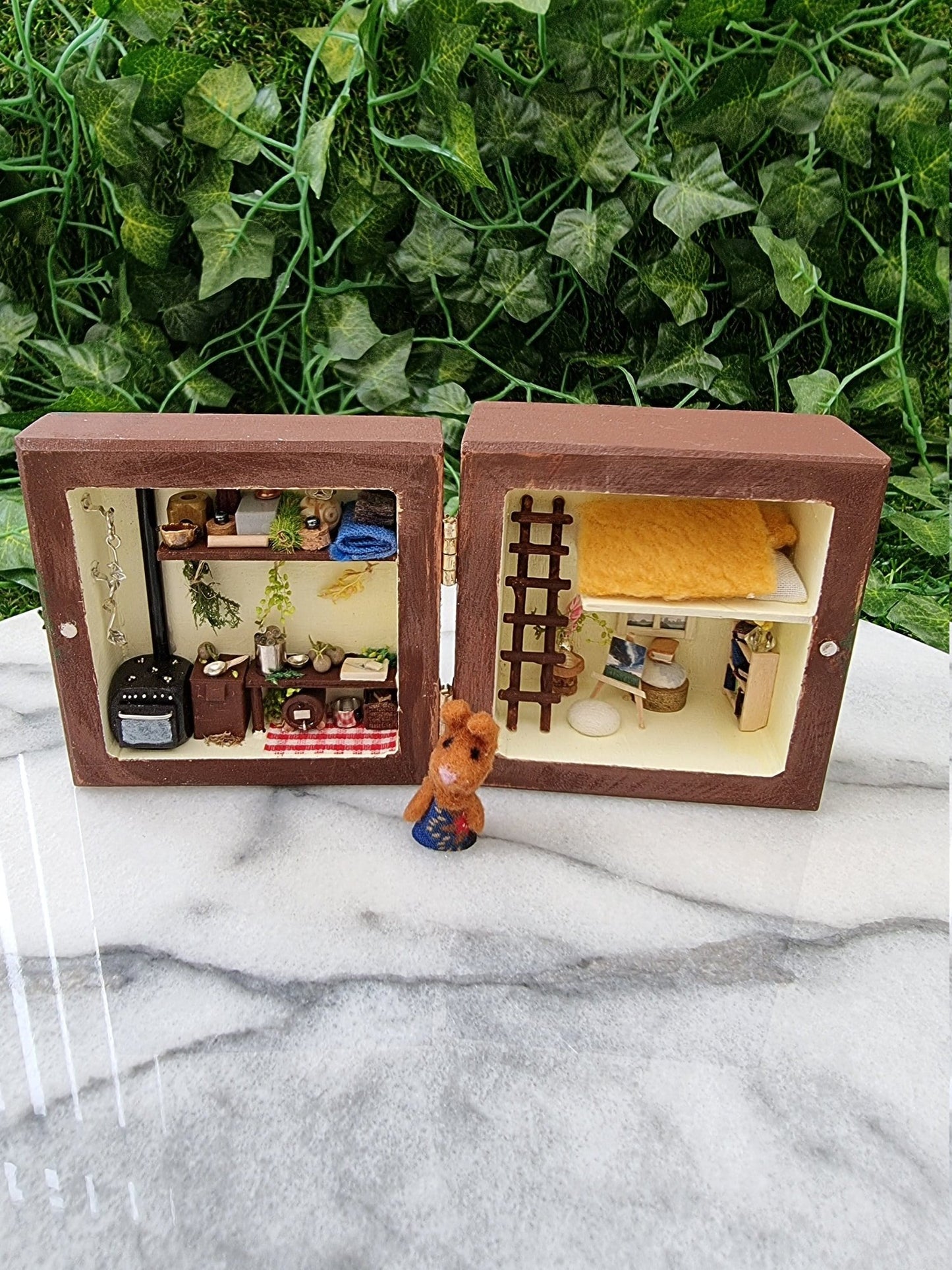 A small brown box sits open like a book on a marble slab. A small brown bunny in a dress sits in front, with the left side showing a tiny oven, table, keg, herbs and storage. The right side has a bookcase, easel and a ladder up to a loft bed