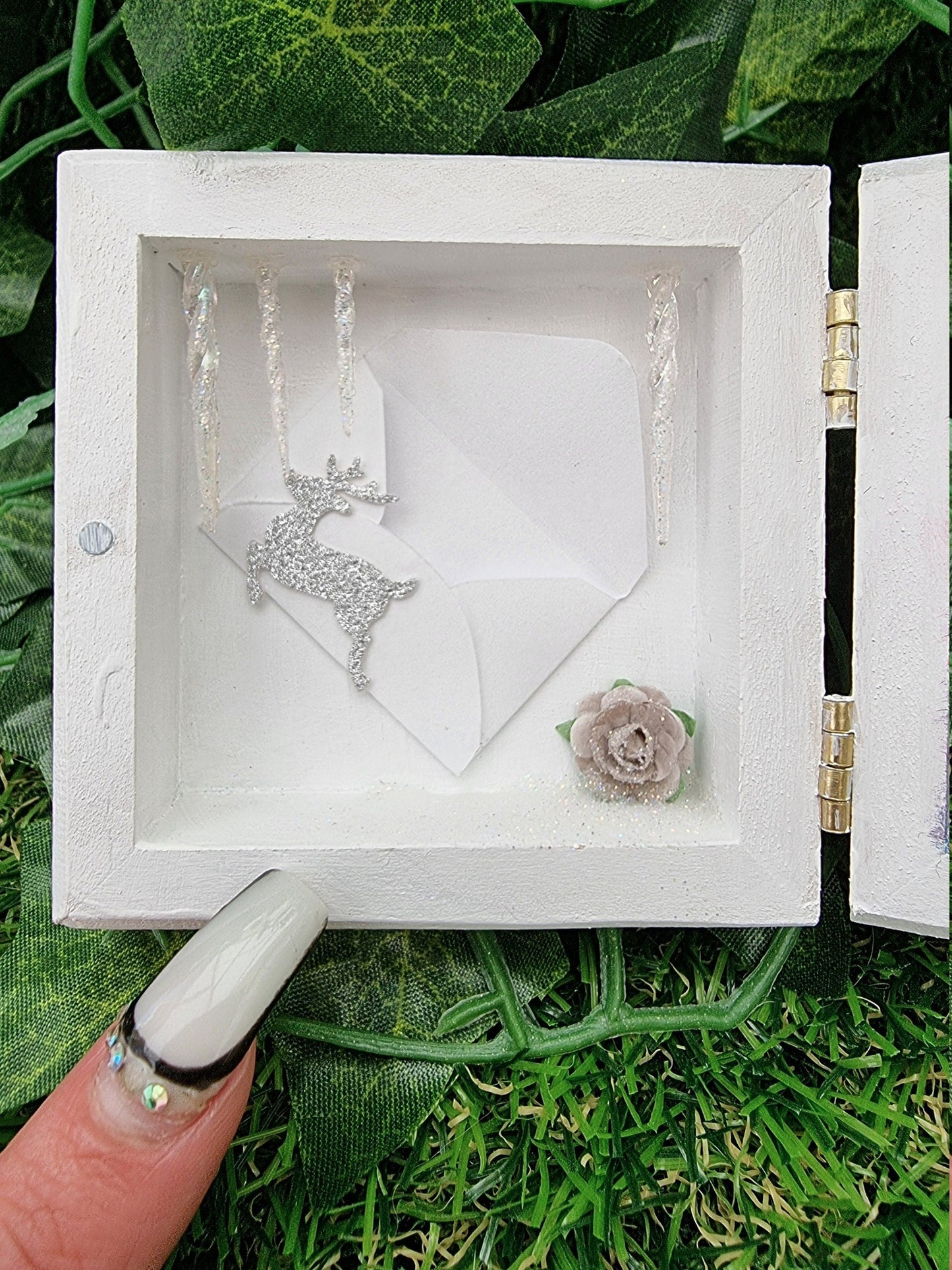A small white square box with lid, open like a book. Detail shown of icicles hanging down from the top. A miniature envelope with a reindeer motif in silver glitter is placed on the back wall. A grey rose is bottom right. A finger points for scale