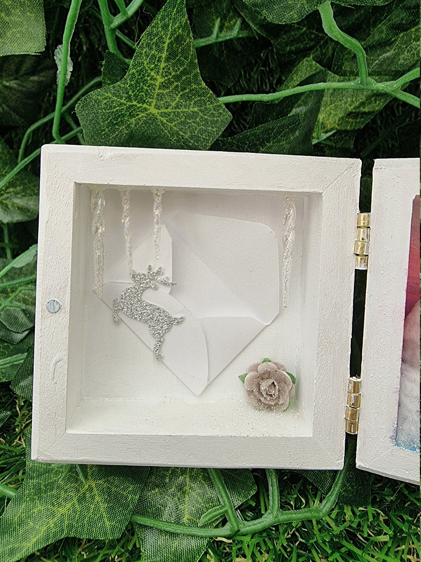 A small white square box with lid, open like a book. Detail is shown of the left side, which has icicles hanging down from the top. A miniature envelope with a reindeer motif in silver glitter is placed on the back wall. A grey rose is bottom right