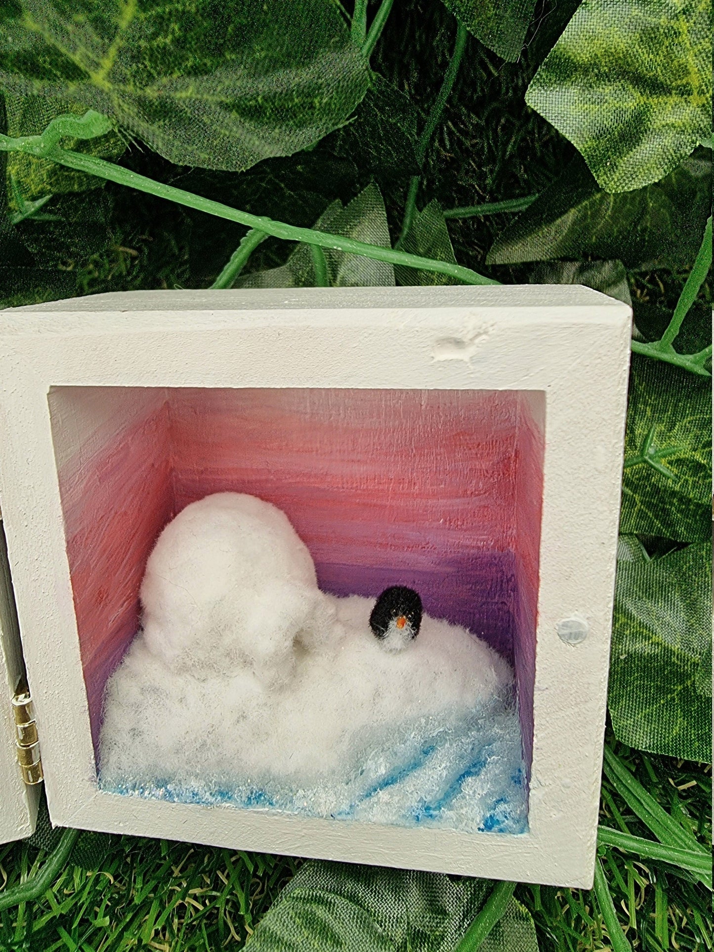 A small white square box, open like a book. Detail is shown of the scene on the right, showing a purple to pink ombre sunset, turquoise and white sea surrounding a snowy iceberg with a white igloo and a tiny penguin.
