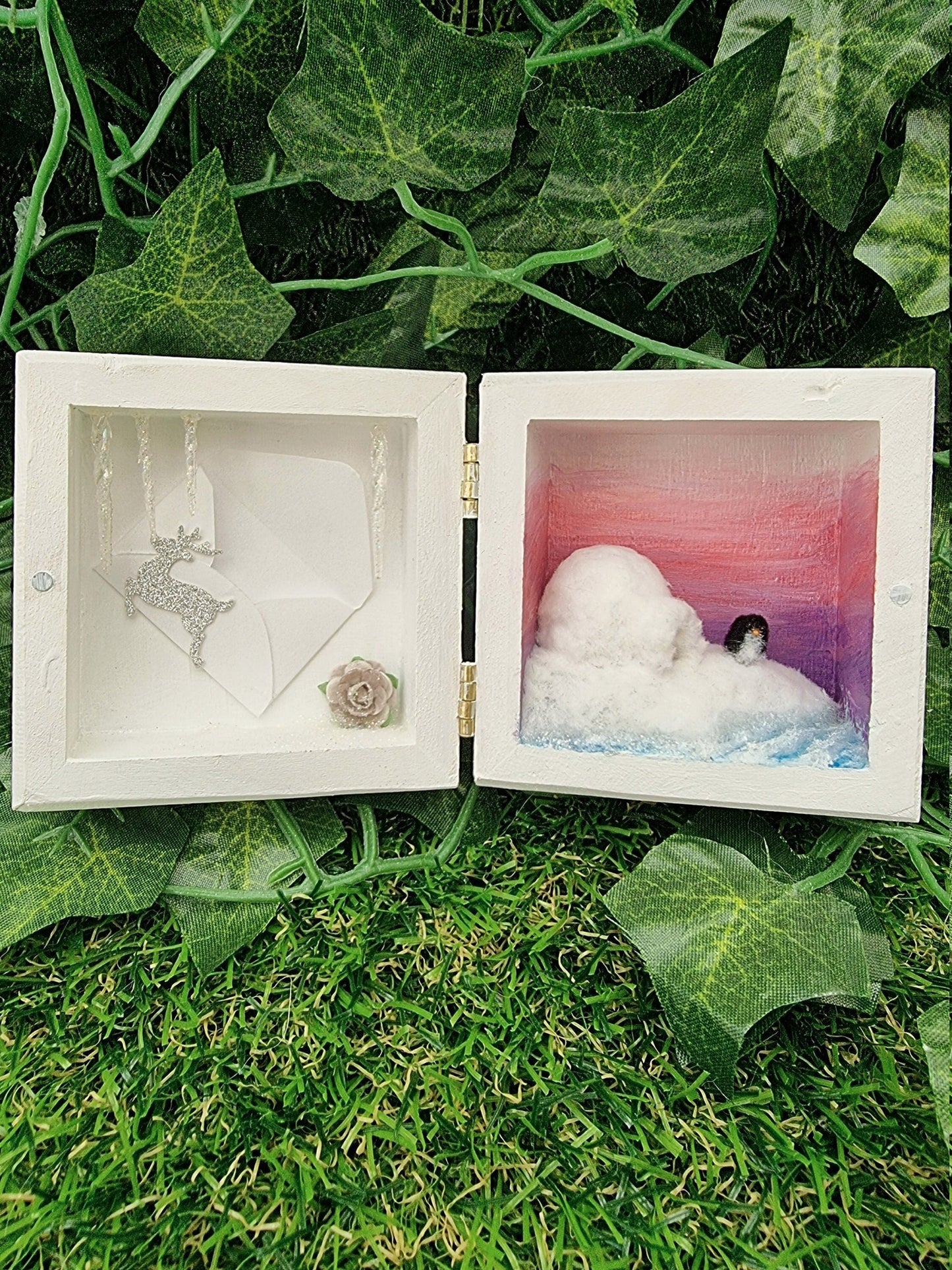 A small white square box, open like a book. On the left, the box has icicles, miniature envelope, grey rose and a reindeer motif in silver glitter. On the right, the box contains a diorama with pink sunset, blue sea, white igloo and a tiny penguin.