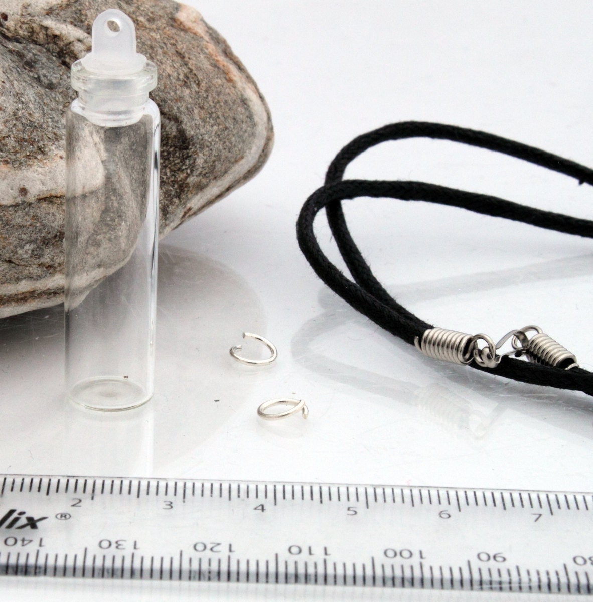 Tall glass vial with a plastic lid sits beside a ruler, jump rings and a black pendant cord