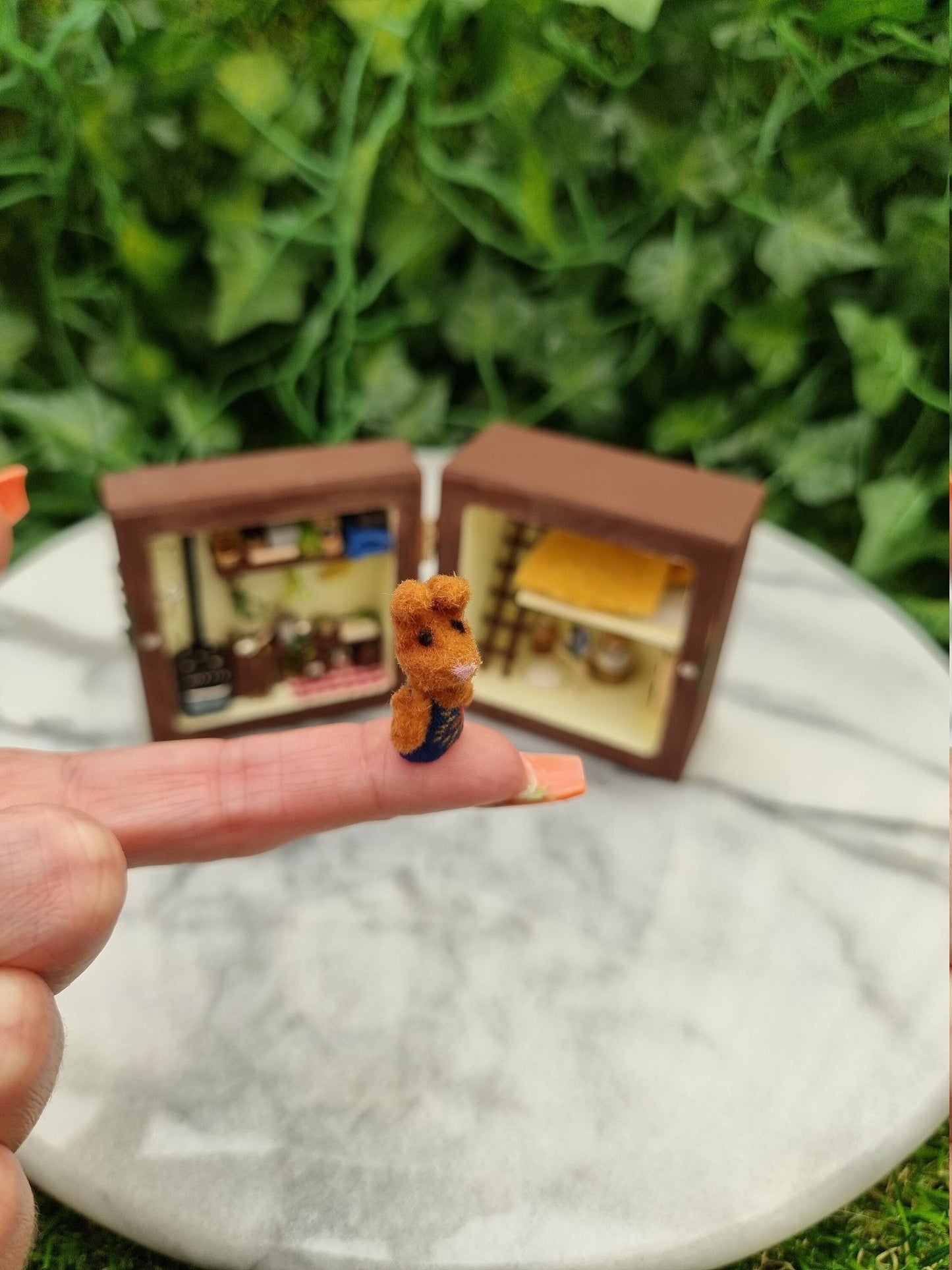 A small brown bunny in a dress sits on a finger in front of a small brown box diorama open like a book in the background with a kitchen, lounge and bedroom