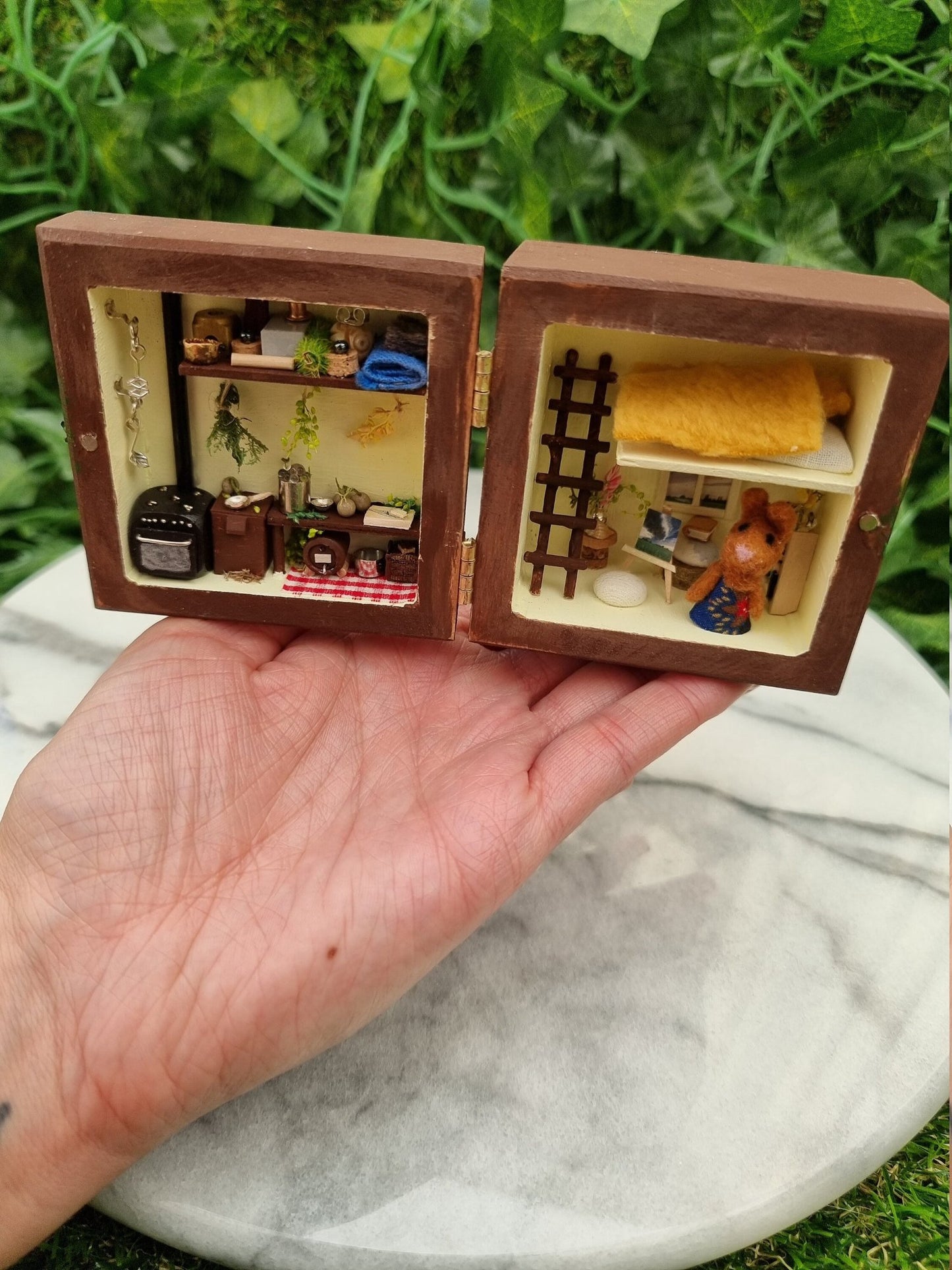 A small brown box sits open like a book on a palm. A small brown bunny in a dress sits in the right side of the box along with a bookcase, table, easel and ladder to a loft bed. The left side has a tiny oven, table, keg, herbs, tools and storage.