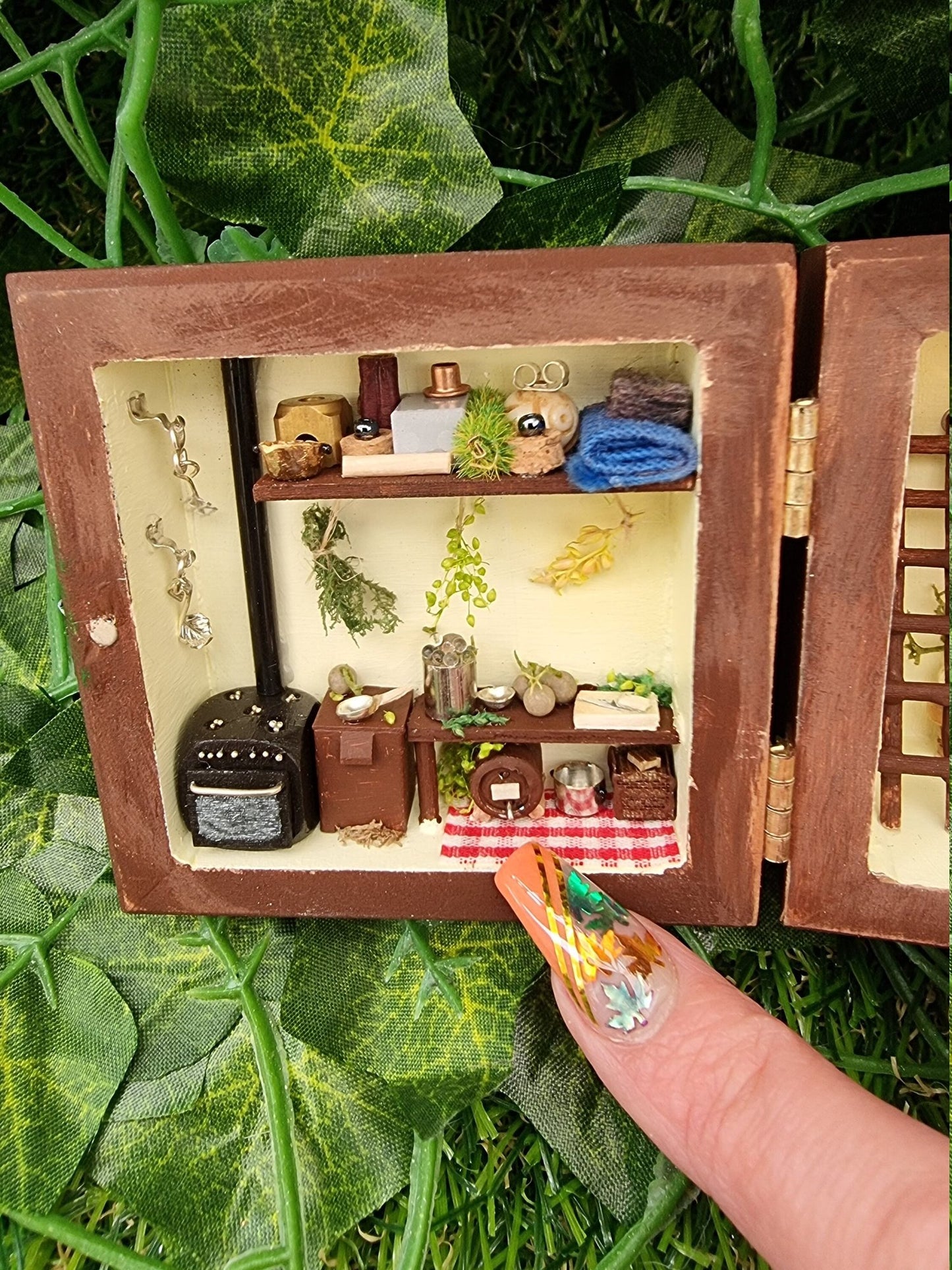 A small brown box sits open like a book. Detail is shown of the left side and a finger points to a ting rug in front of a keg, cooking pot and crate. There is a table with food being prepared, a tiny oven, herbs, tools, blankets and storage.