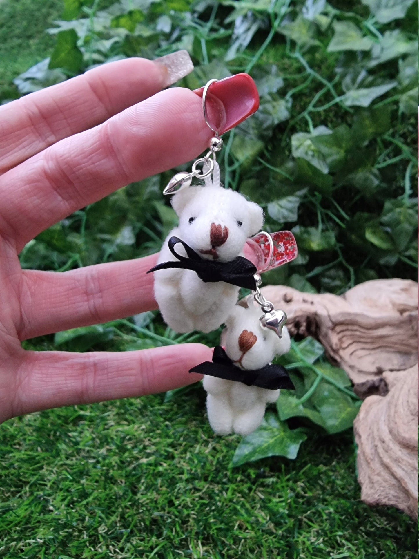 Hand holds two silver coloured hook style earrings, each with a small silver coloured fluffy heart charm and cream coloured fabric teddy bear. Each teddy bear wears a black ribbon bow tie