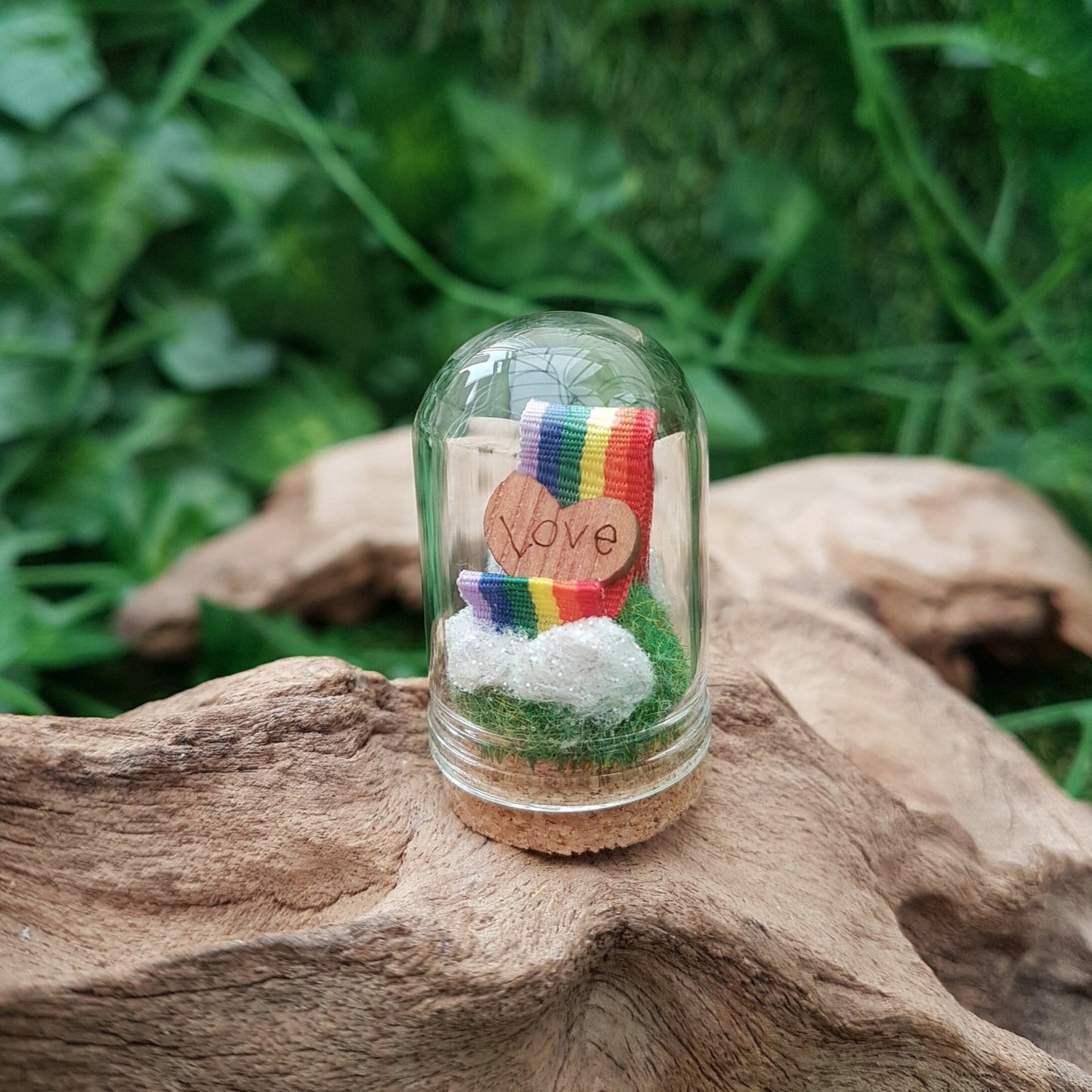 Miniature glass bell jar containing a curl of rainbow ribbon with miniature clouds at the start and end of the ribbon at the back and front. A small wooden heart with sits on the ribbon, engraved with the word LOVE