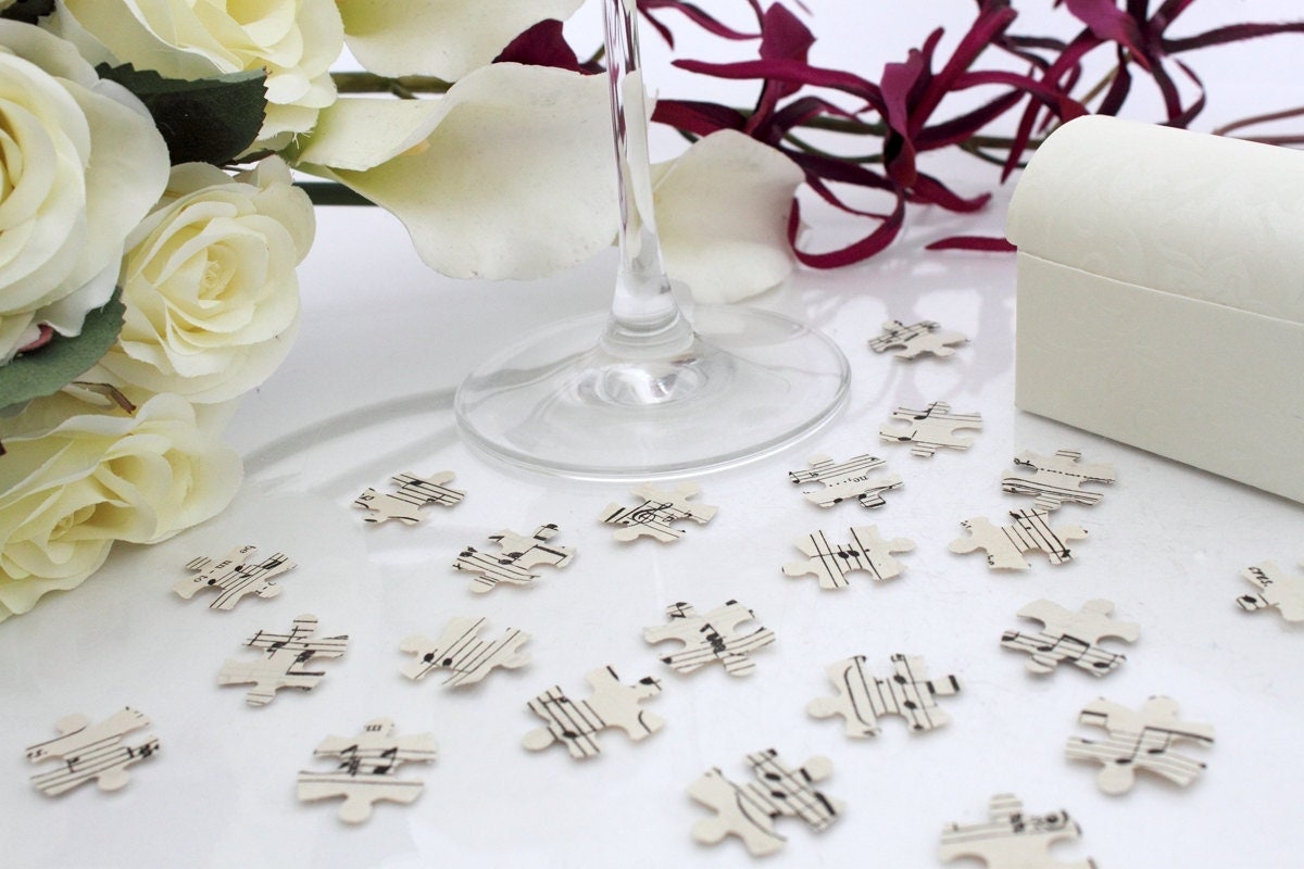 Cream paper confetti pieces in jigsaw shapes are scattered on a white background with a champagne flute and purple orchid bouquet in the background. The paper confetti is cut from sheet music