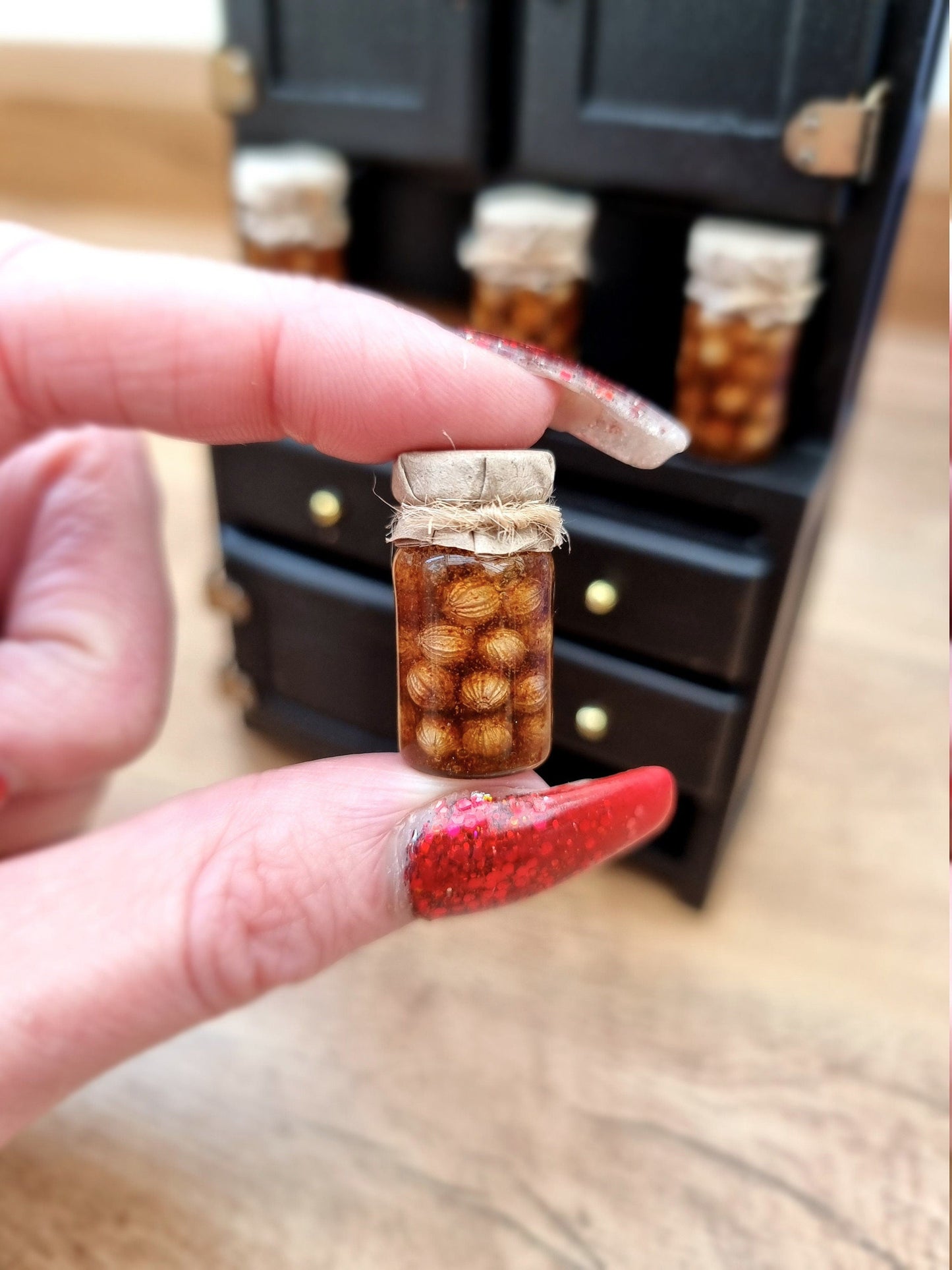 Single 1:12 scale miniature preserve jar with paper lid containing pickled onions. The jar is held between thumb and finger. There is a miniature food cabinet in the background