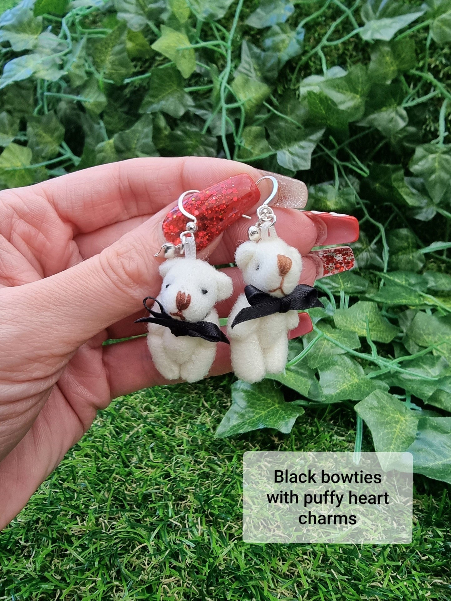 Hand holds two silver coloured hook style earrings, each with a small silver coloured fluffy heart charm and cream coloured fabric teddy bear. Each teddy bear wears a black ribbon bow tie