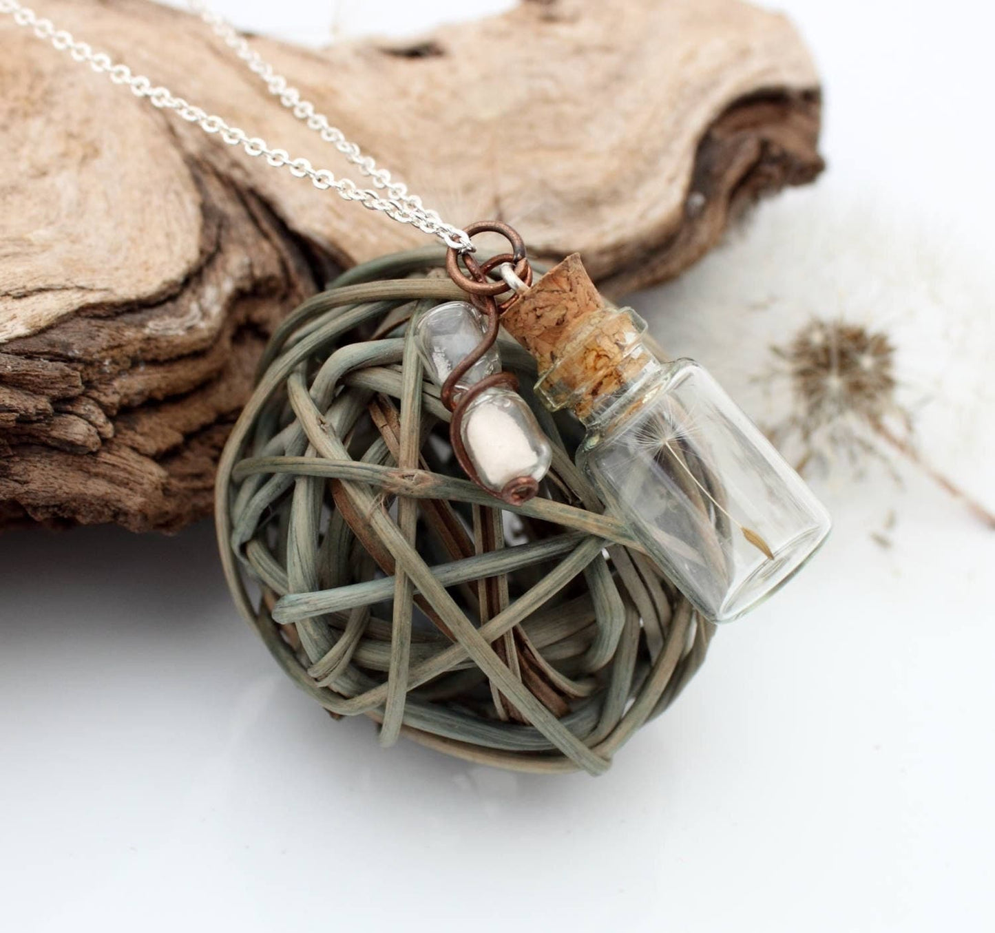 Miniature jar shaped glass vial necklace containing a dandelion seed with a copper wrapped miniature glass sand timer attachdd to the cork lid. Displayed on a silver coloured chain attached to the cork lid