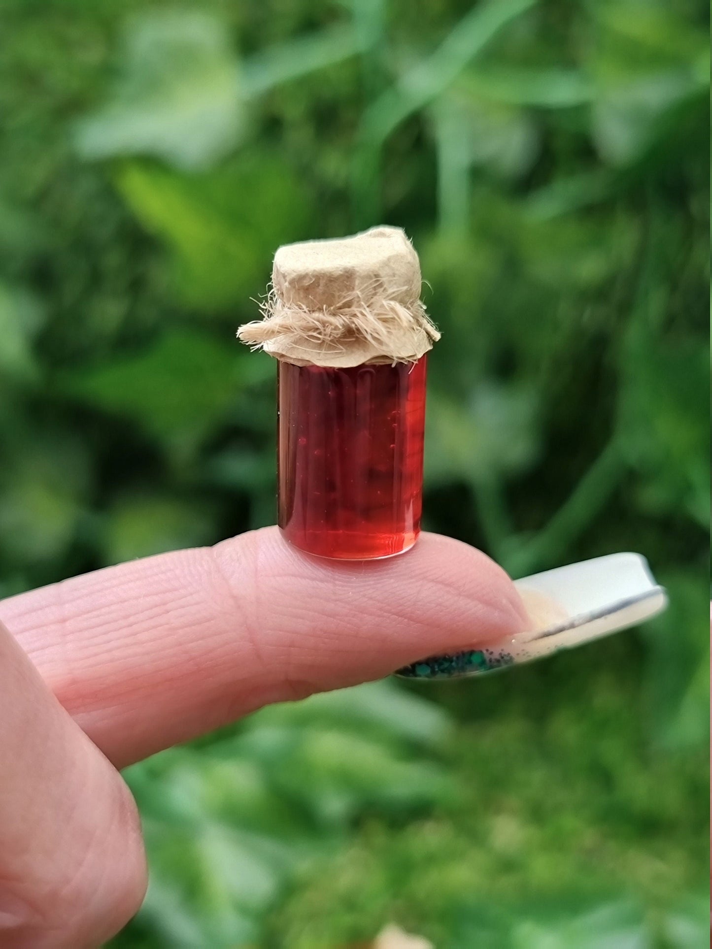A miniature jar of crimson jam with brown paper lid is shown sat on an index finger against a green background