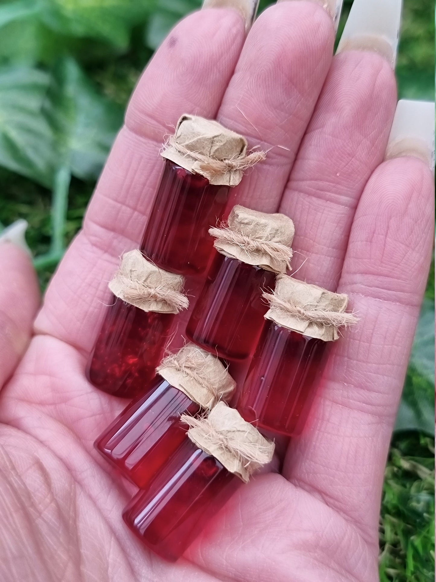 Several miniature jars of crimson jam with brown paper lids are show held in the palm of a hand against a green background