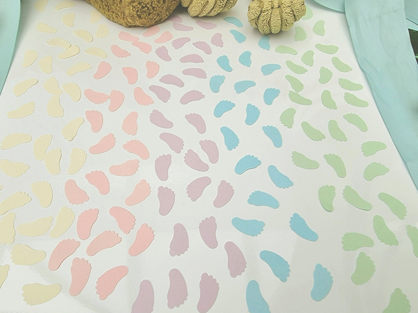 Blocks of pastel cream, pink, lilac, blue and green baby feet shaped confetti pieces are scattered on a white background. Cuddly toys are seen in the background