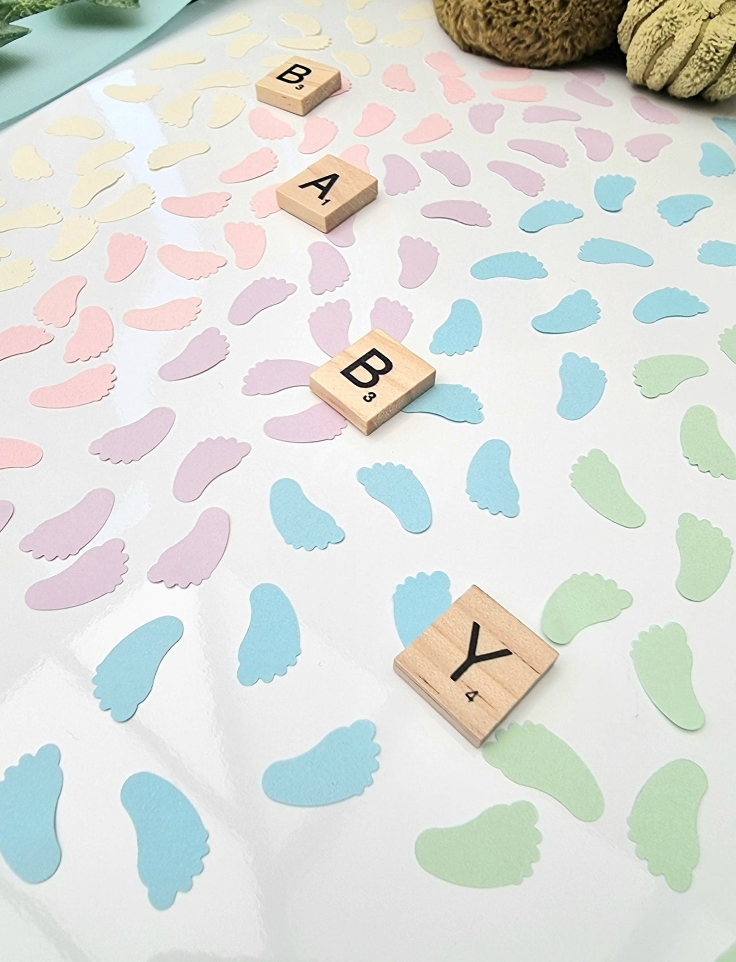 Blocks of pastel cream, pink, lilac, blue and green baby feet shaped confetti pieces are scattered on a white background with wooden scrabble tiles reading BABY diagonally across the photo. Cuddly toys are seen in the background