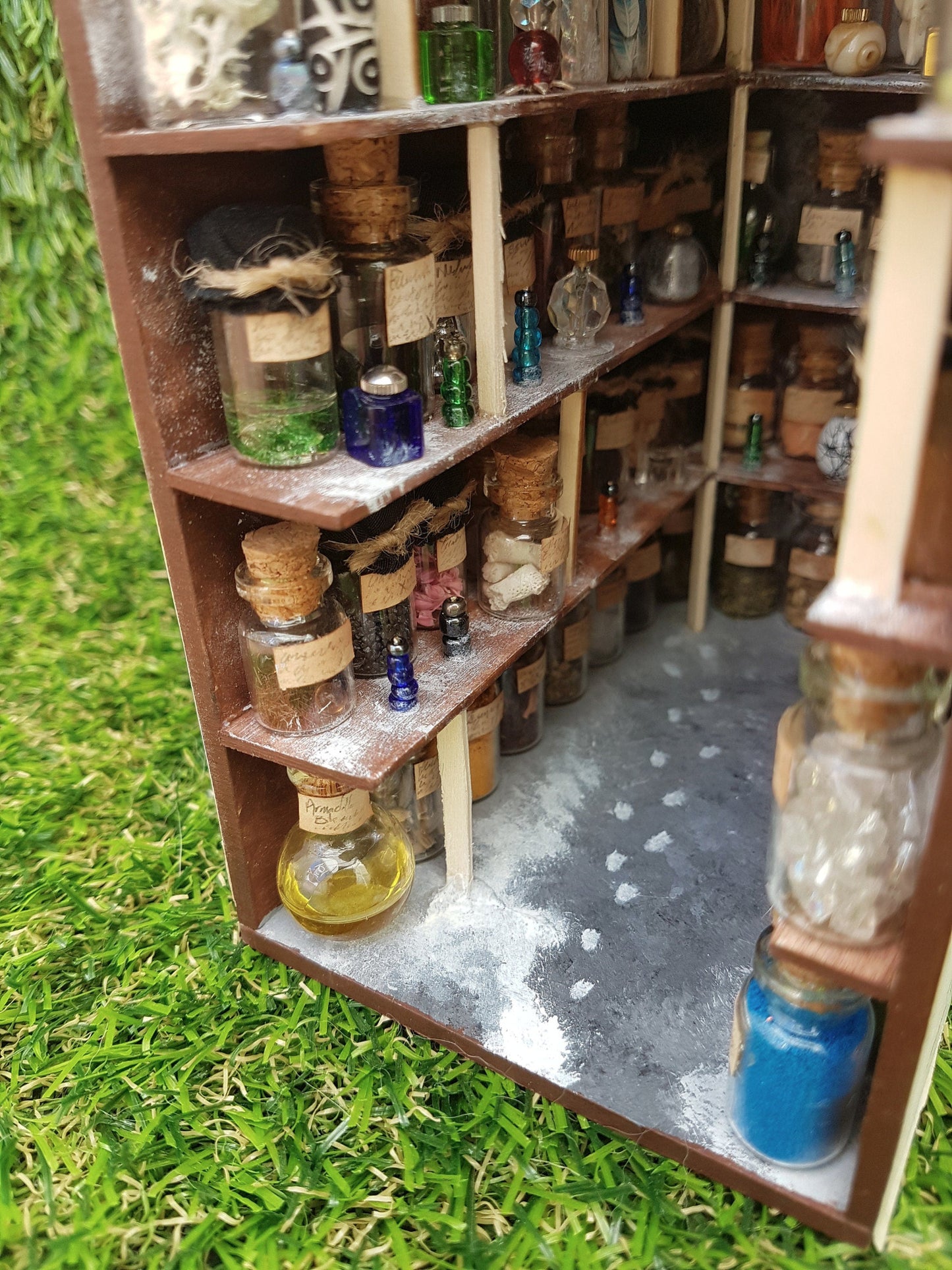 Photo looks down onto the floor of the potions store and 3 shelves. The shelves are filled with glass bottles and vessels. The floor is grey and shows a spillage of something white. There are white footprints on the floor leading to the back