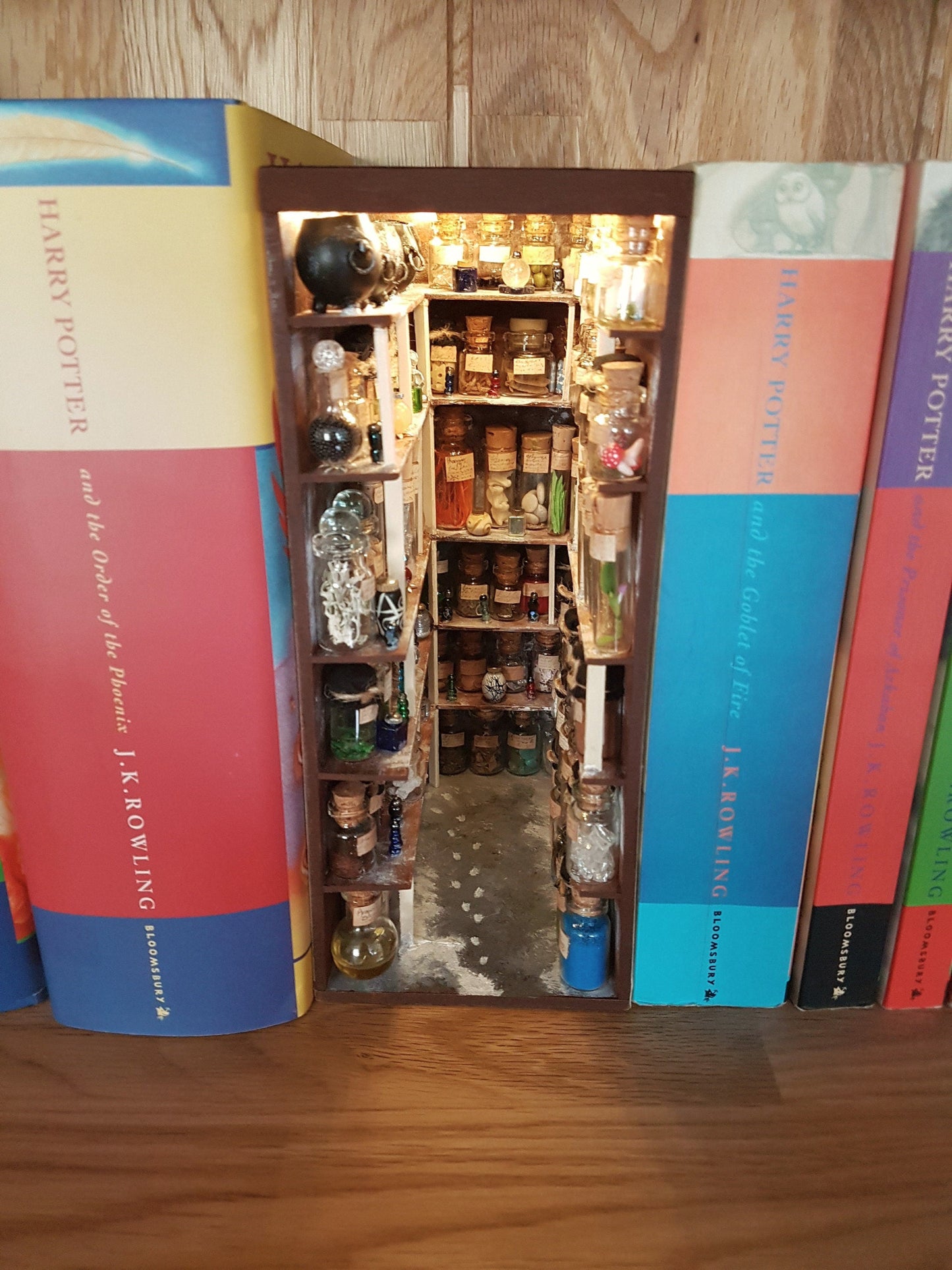 A small open fronted book nook shelf insert box sits between books of the same size on a bookshelf. The box contains 5 sets of shelves up to the ceiling on all sides, each full of glass vials and vessels. A row of cauldrons is seen in the top left.