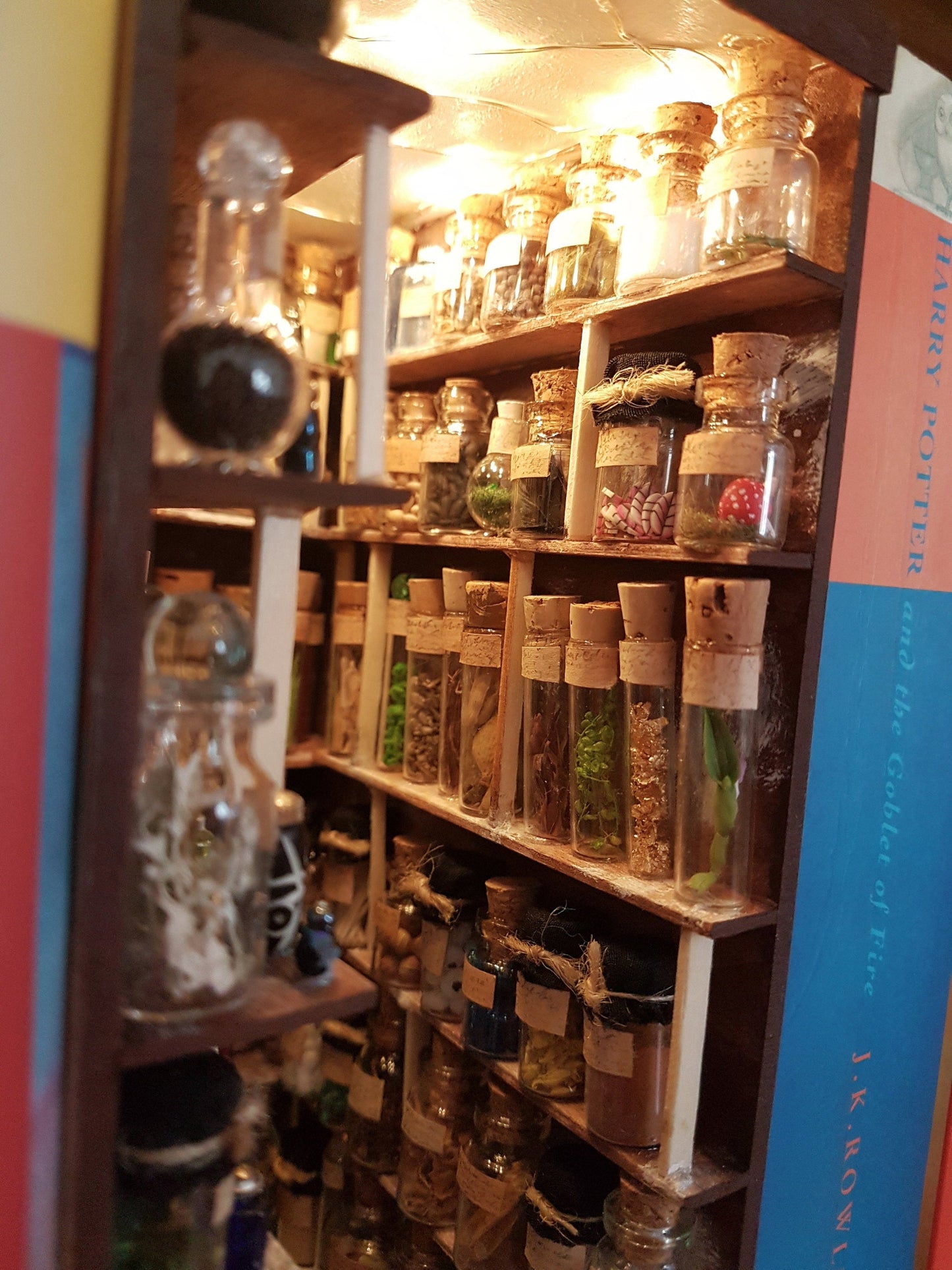 A small open fronted book nook shelf insert box sits between books of the same size on a bookshelf. The box contains 5 sets of shelves up to the ceiling on all sides, full of glass vials and vessels