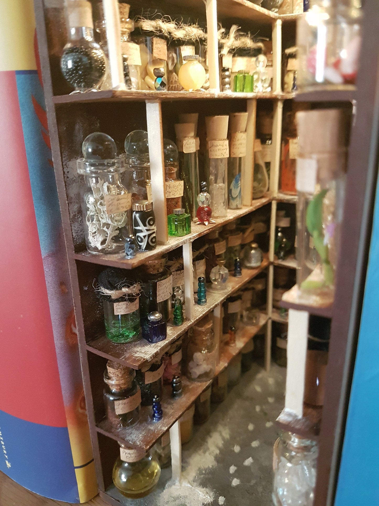 A small open fronted book nook shelf insert box sits between books of the same size on a bookshelf. The box contains 5 sets of shelves up to the ceiling on all sides, each full of glass vials and vessels. The booknook is lit with fairy lights