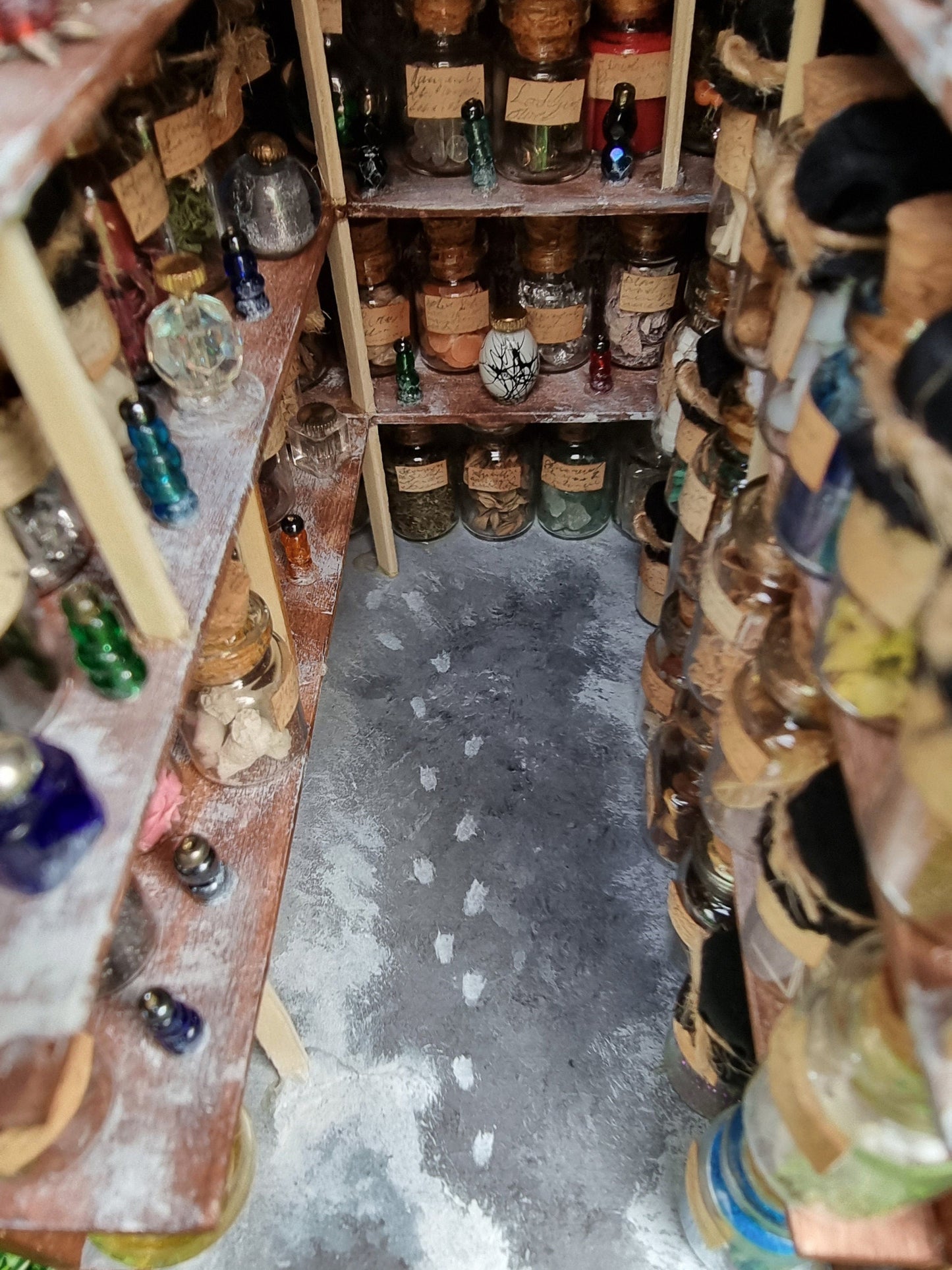 Photo looks down onto the floor of the potions store and 3 shelves. The shelves are filled with glass bottles and vessels. The floor is grey and shows a spillage of something white. There are white footprints on the floor leading to the back