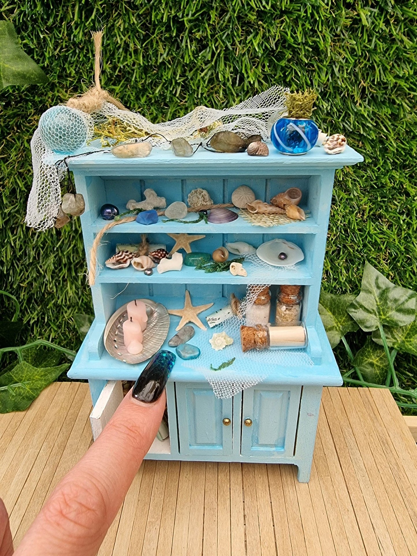 A 1:12 miniature pastel blue dresser sits on a wooden floor against a green background. The dresser displays glass vials, netting, glass floats, shells and candles in a shell dish. A finger points at the dresser for scale