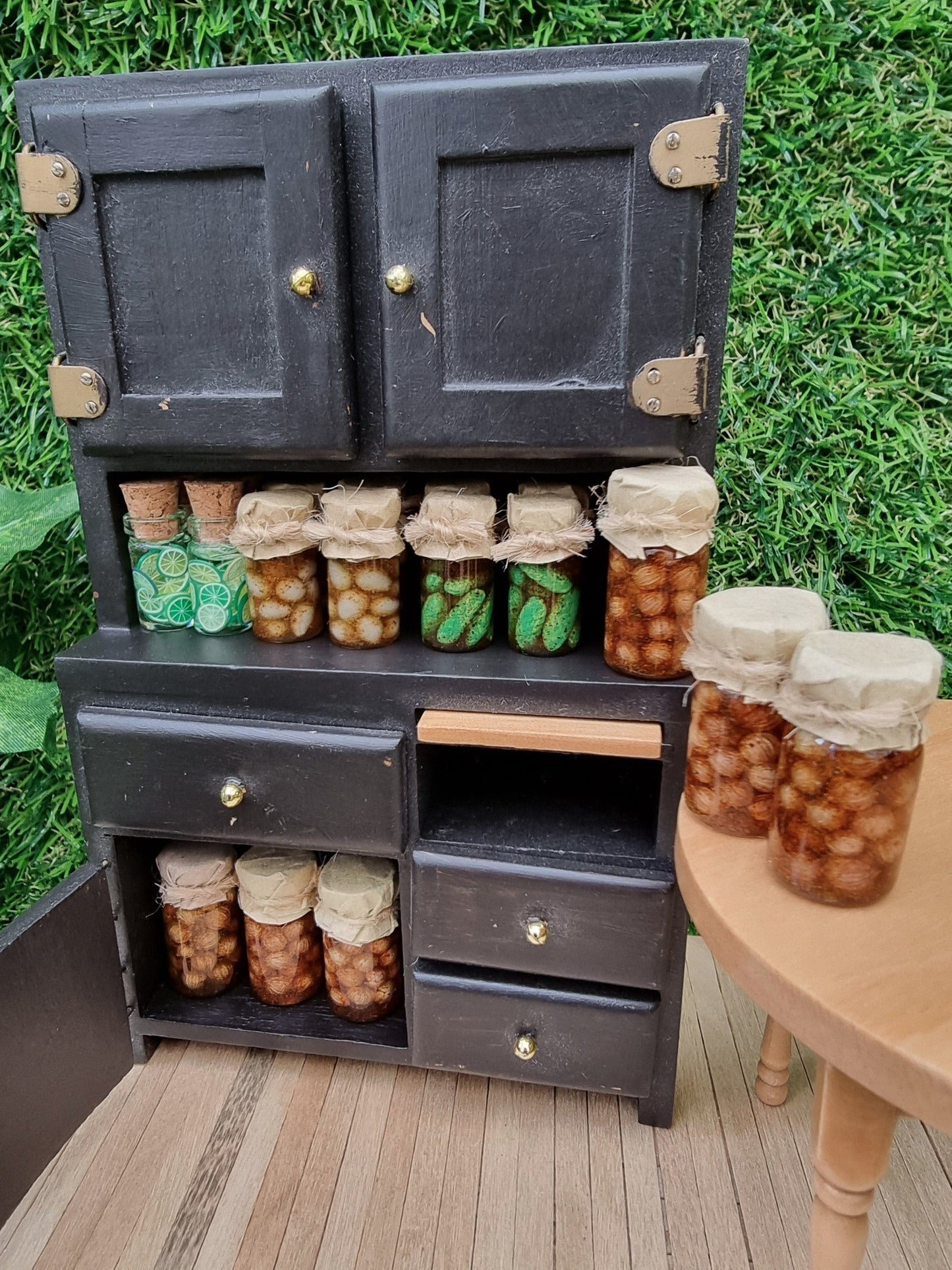 Miniature 1:12 scale black food cupboard containing several miniature preserve jars with paper lidss. Jars contain pickled onions, pickled eggs, gherkins and lime slices
