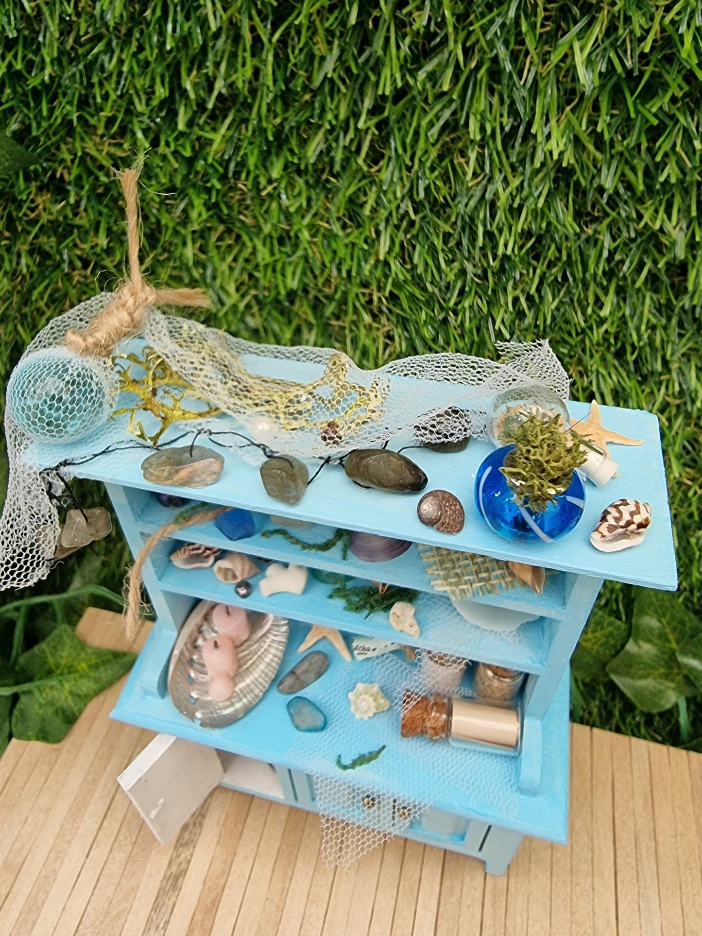 A 1:12 miniature pastel blue dresser pictured from above sits on a wooden floor. The dresser displays glass vials, netting, glass floats, shells and candles in a shell dish. The dresser had 3 cupboard doors. One drawer is open