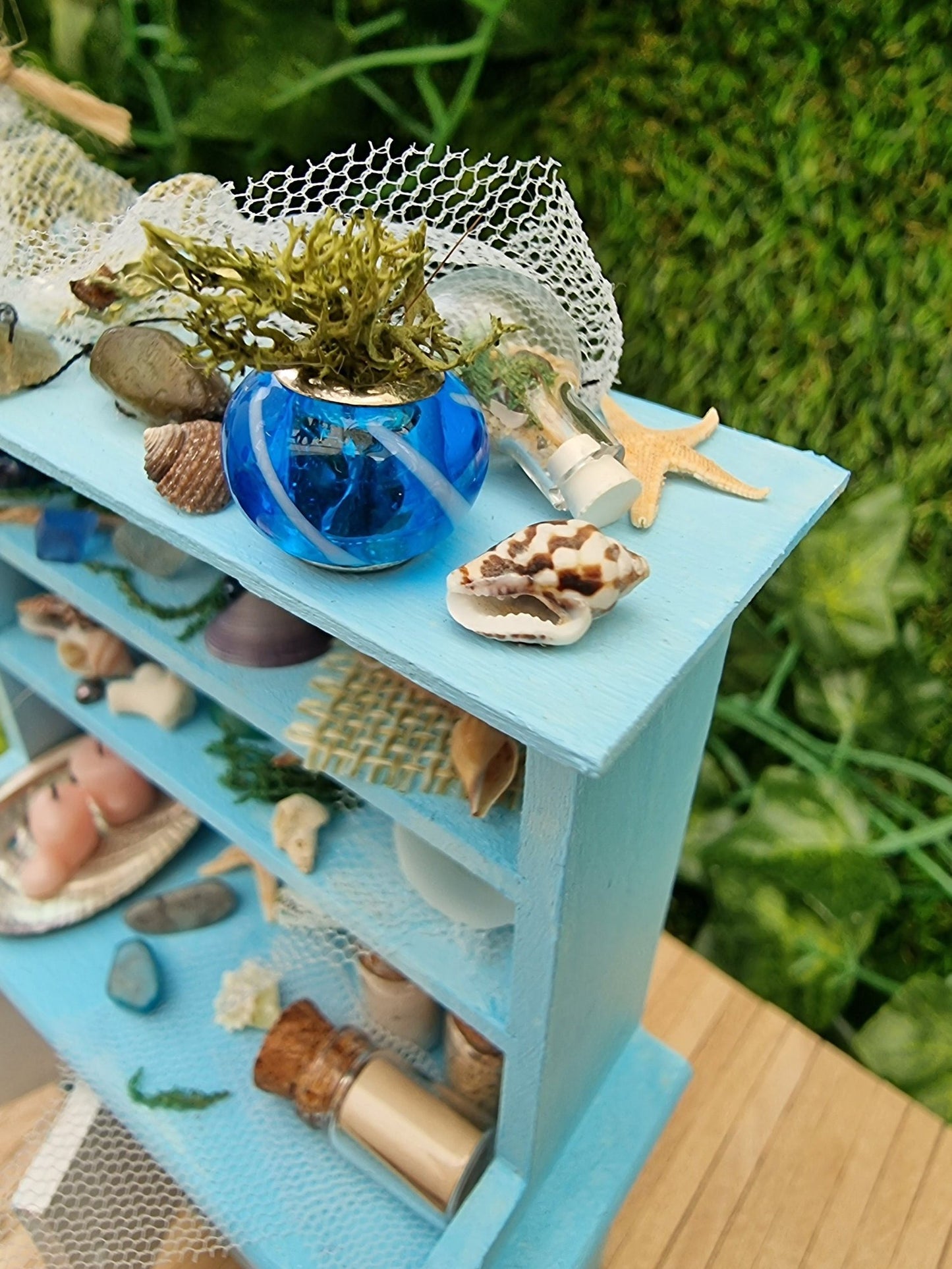 A 1:12 miniature pastel blue dresser sits on a wooden floor against a green background. The dresser displays glass vials, netting, glass floats, shells and candles in a shell dish. A blue vase sits on top