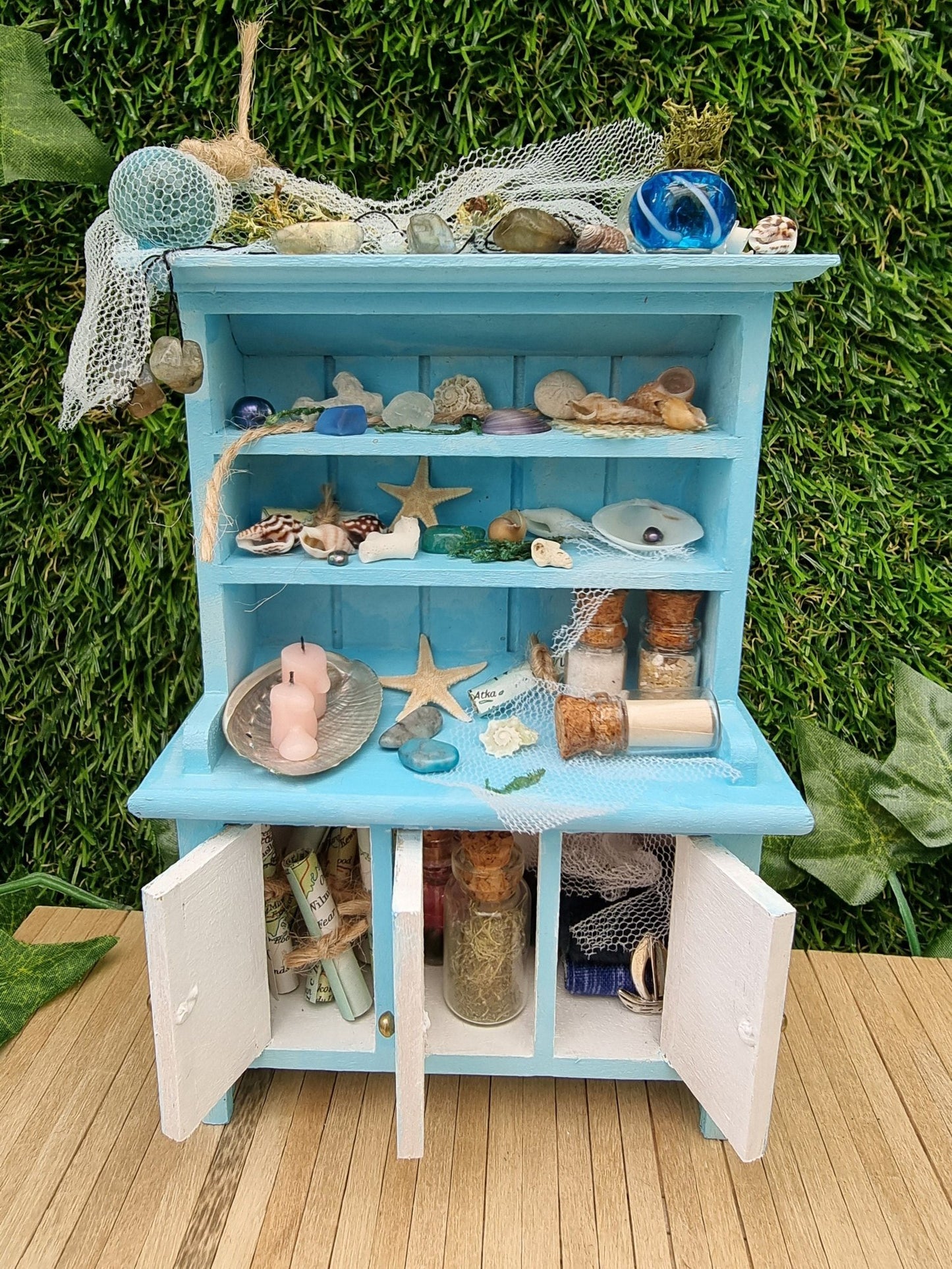 A miniature pastel blue dresser sits on a wooden floor. The dresser displays glass vials, netting, glass floats, starfish, shells and candles in a shell dish. The dresser has 3 open cupboard doors revealing maps and glass bottles inside