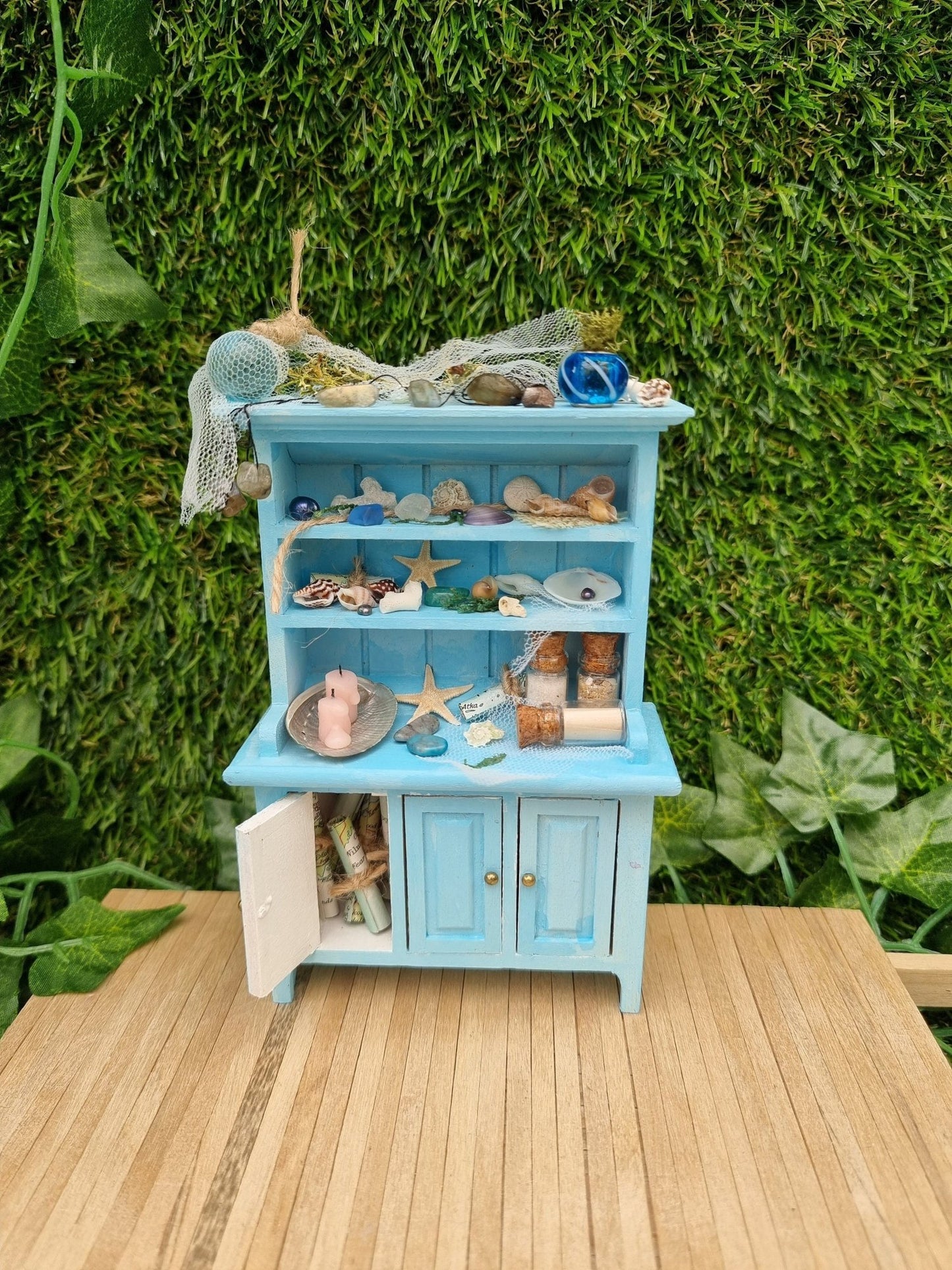 A miniature pastel blue dresser sits on a wooden floor against a green background. The dresser displays glass vials, netting, glass floats, shells and candles in a shell dish. The dresser had 3 cupboard doors. One is open revealing maps inside