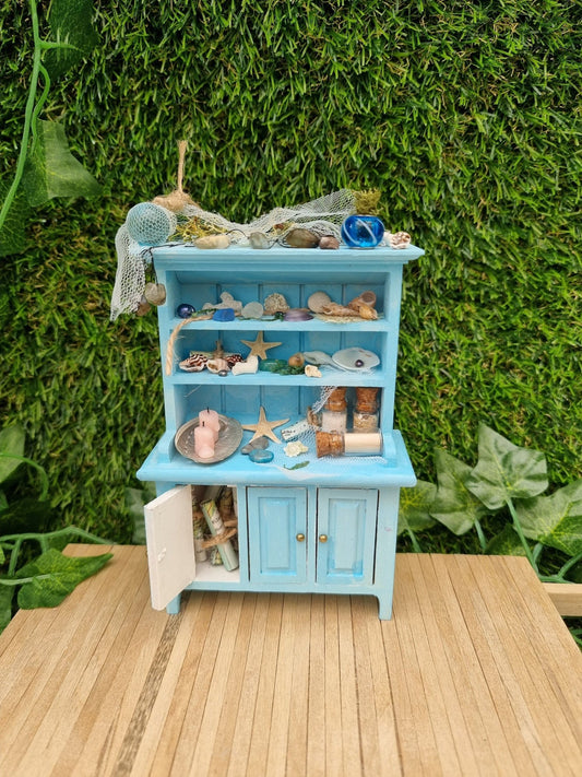 A miniature pastel blue dresser sits on a wooden floor against a green background. The dresser displays glass vials, netting, glass floats, shells and candles in a shell dish. The dresser had 3 cupboard doors. One is open revealing maps inside