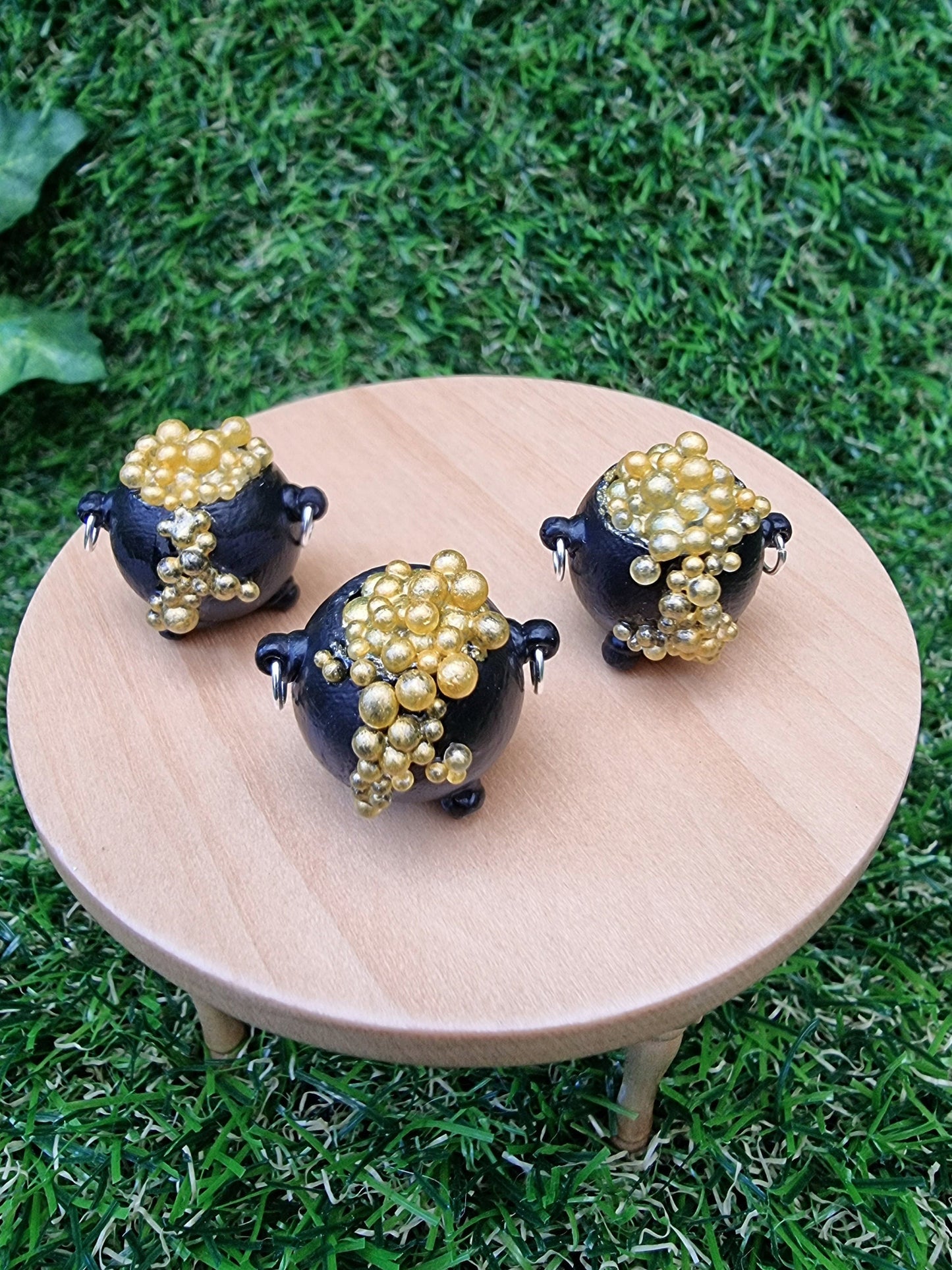 3 miniature black cauldrons overflowing with golden bubbles are displayed on a miniature wooden table against a green grass background