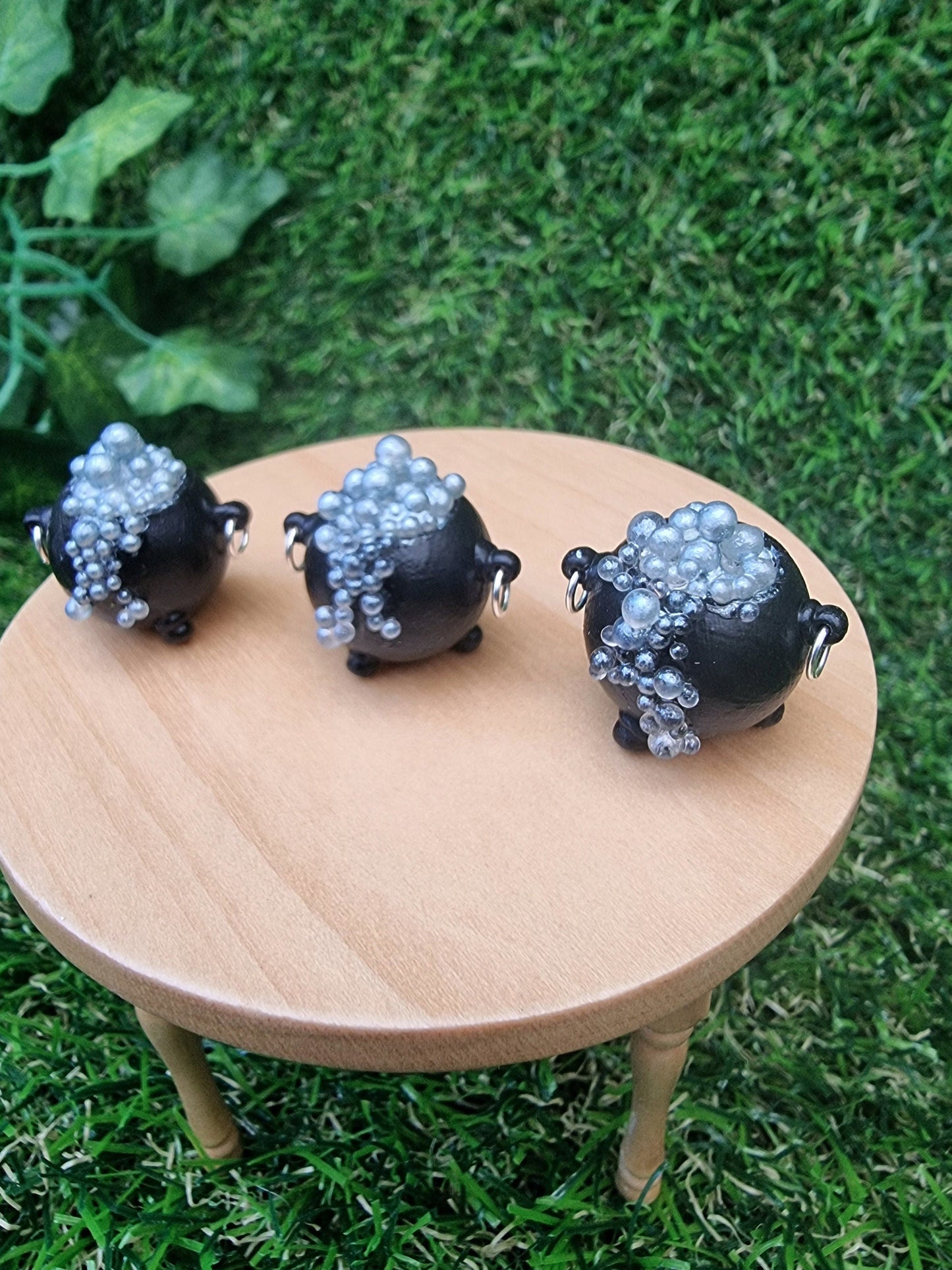 3 miniature black cauldrons overflowing with silver coloured bubbles are displayed on a miniature wooden table against a green grass background