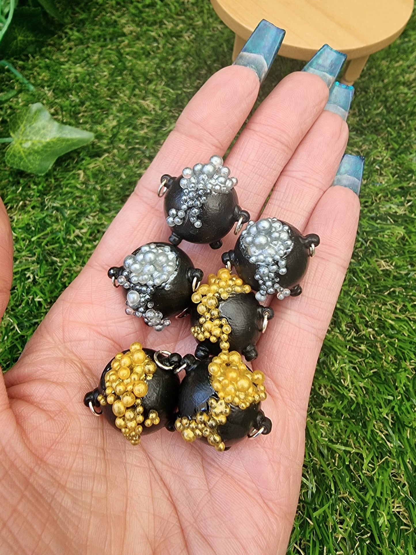 6 miniature black cauldrons overflowing with bubbles are displayed in an open palm. 3 of the cauldrons have bubbles in a gold colour. The other 3 have bubbles in silver colour