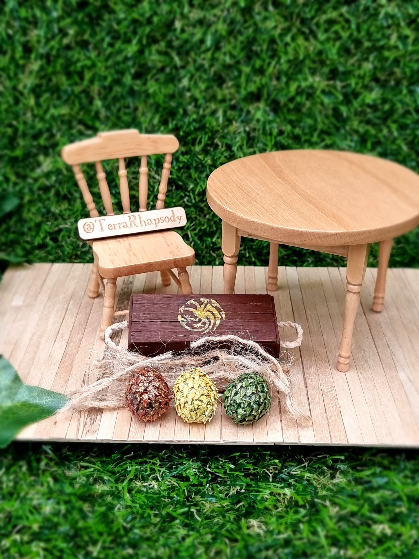 A miniature brown crate and lid are shown with 3 scaled dragon eggs- one red, one green and one yellow, all with a gold sheen. The crate lid shows a gold Targaryen 3-headed dragon crest. The crate has rope handles on the short edge.