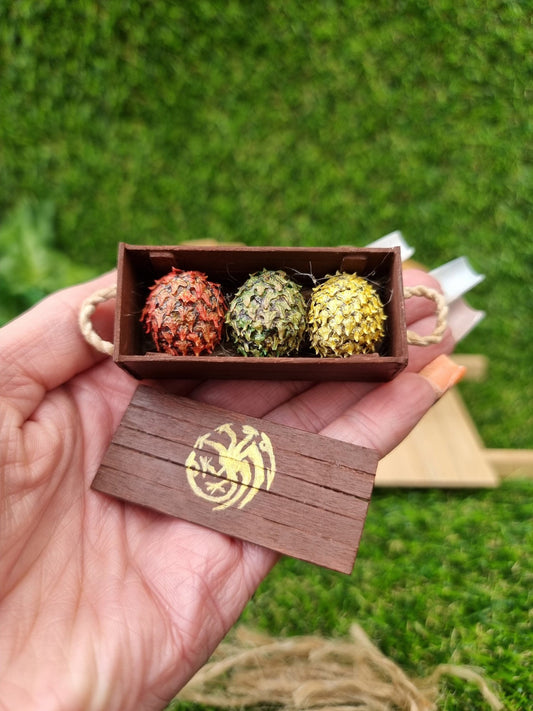 A hand holds a miniature brown crate and lid. The crate contains 3 scaled dragon eggs- one red, one green and one yellow, all with a gold sheen. The crate lid shows a gold Targaryen 3-headed dragon crest. The crate has rope handles oh the short edge