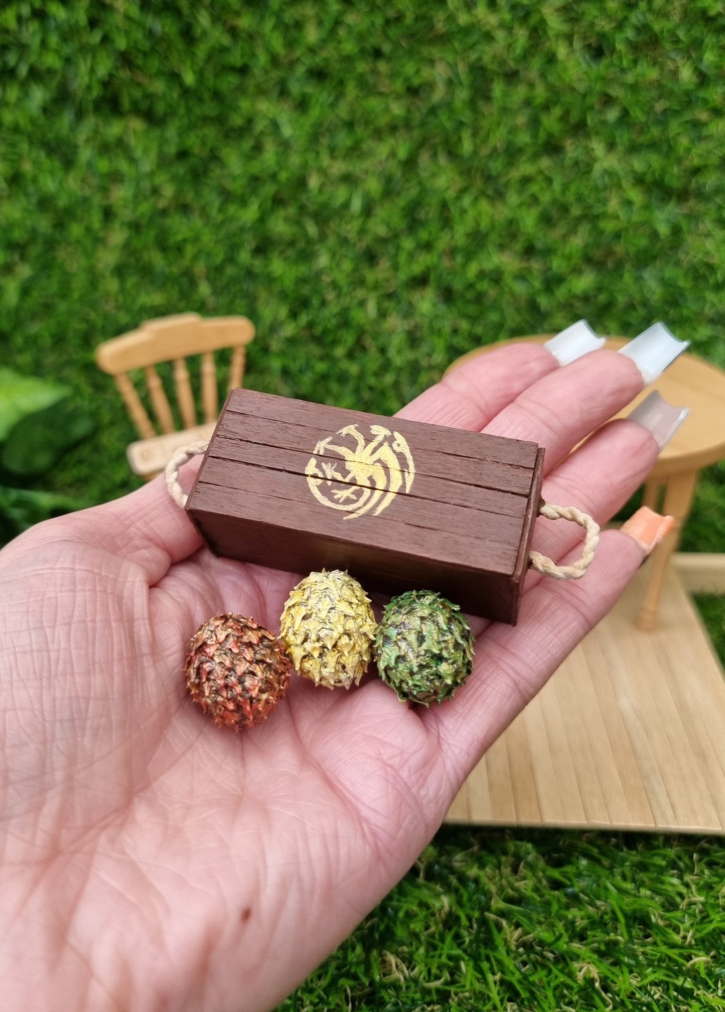 A hand holds a miniature brown lidded crate and 3 scaled dragon eggs- one red, one green and one yellow, all with a gold sheen. The crate lid shows a gold Targaryen 3-headed dragon crest. The crate has rope handles on the short edge